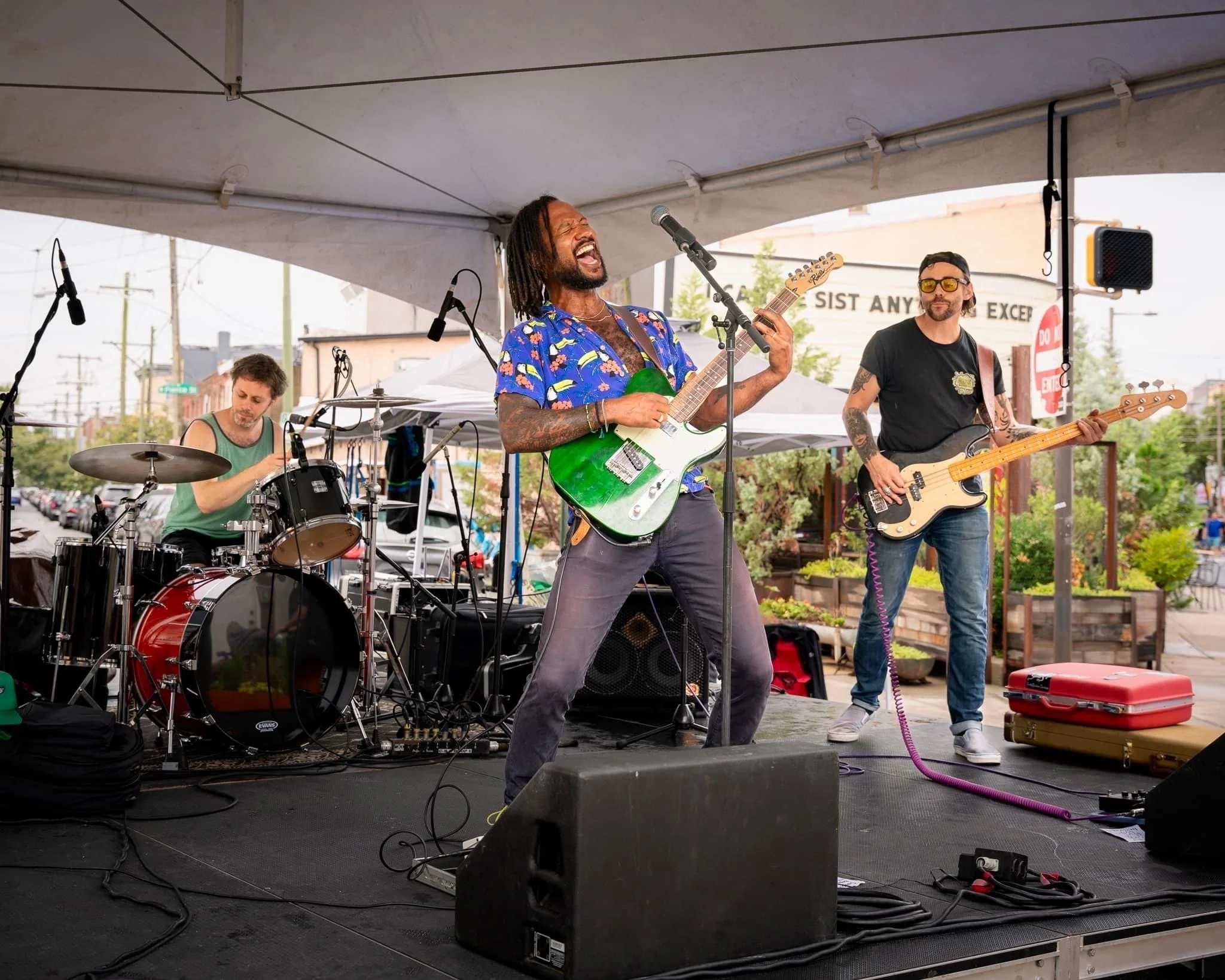 A band performing on an outdoor stage with three musicians: a drummer on the left, a lead guitarist and vocalist center, and a bassist on the right. The lead guitarist is singing passionately and playing a green electric guitar. The scene is set in a