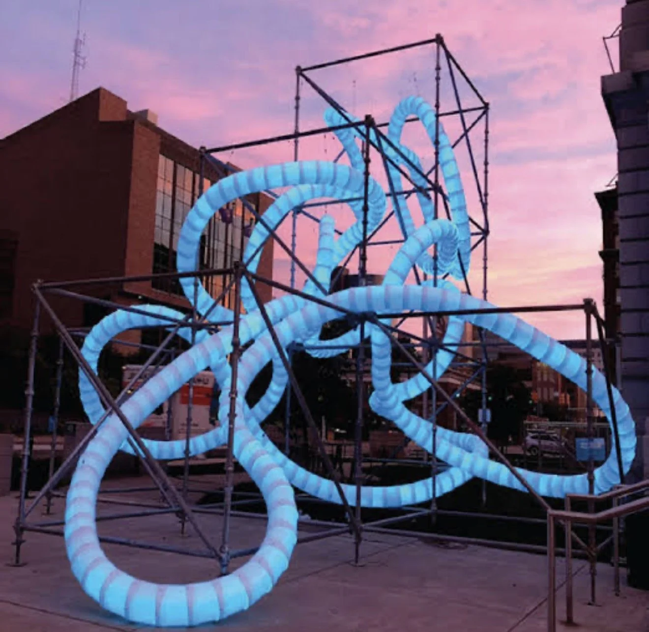 An art installation made of interconnected metal scaffolding with glowing blue tubing, set against a pink and purple sunset sky.