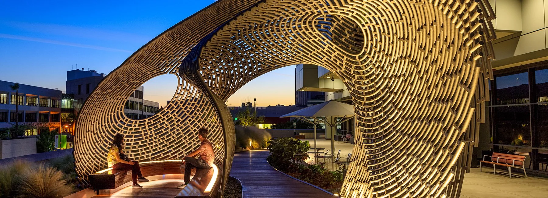 People sitting on a curved bench inside an architecturally intricate lattice structure on a rooftop during dusk.