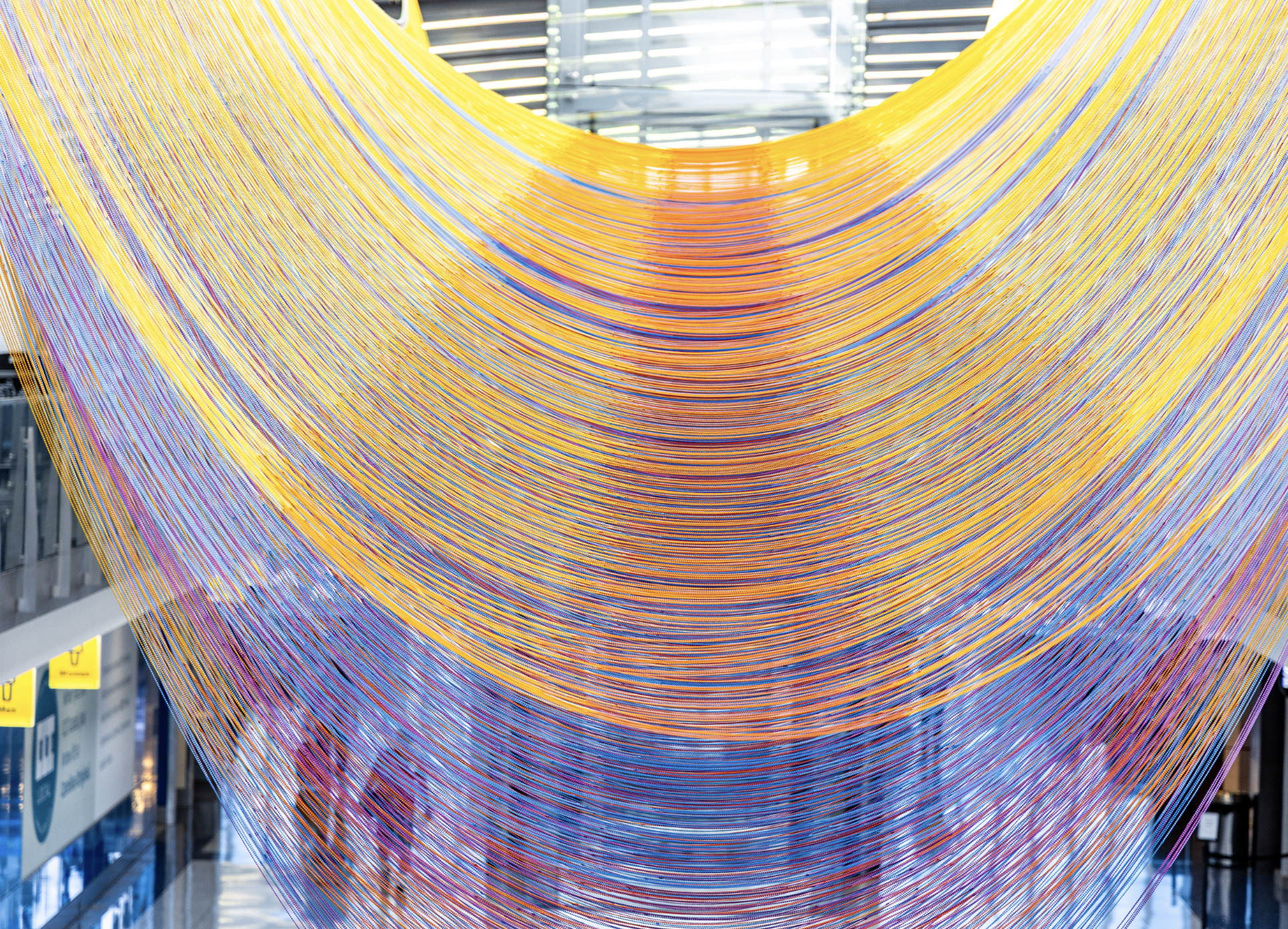 Colorful string art installation with yellow, orange, and purple threads hanging from ceiling in a modern interior space.