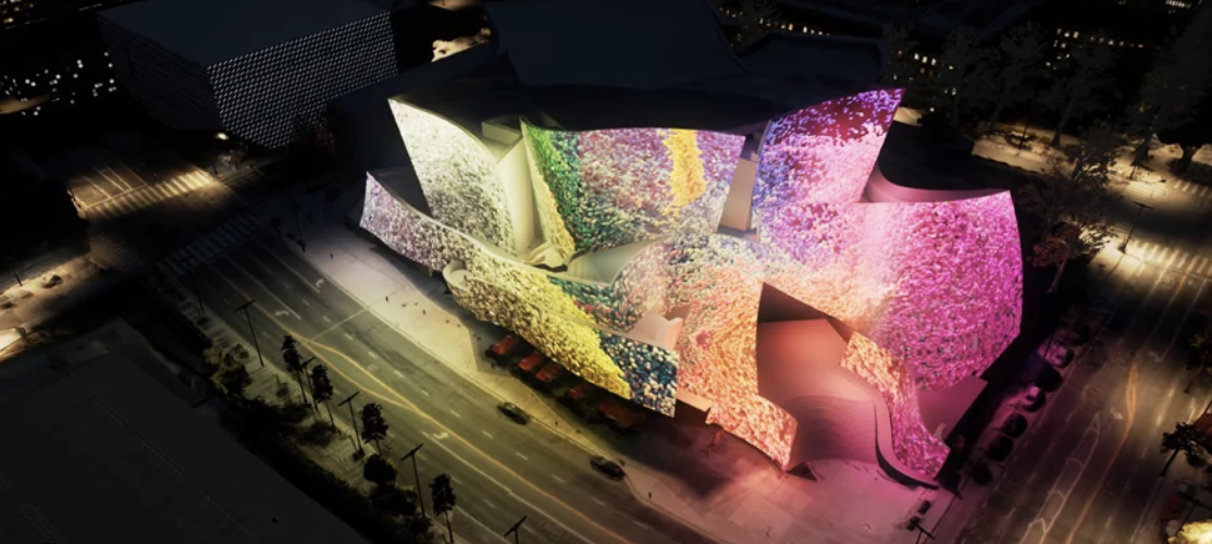 Aerial view of a modern building illuminated with colorful lights, featuring abstract, organic shapes with bright pink, yellow, green, and purple lighting effects, situated along a city street at night.