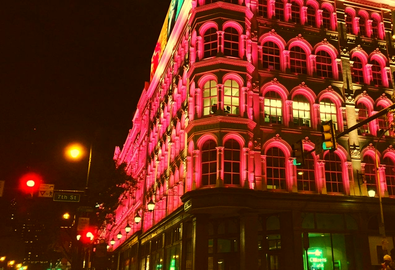 A building at night illuminated with pink lights, decorated with multiple arched windows and ornate architecture, with streetlights and traffic signals in the foreground.