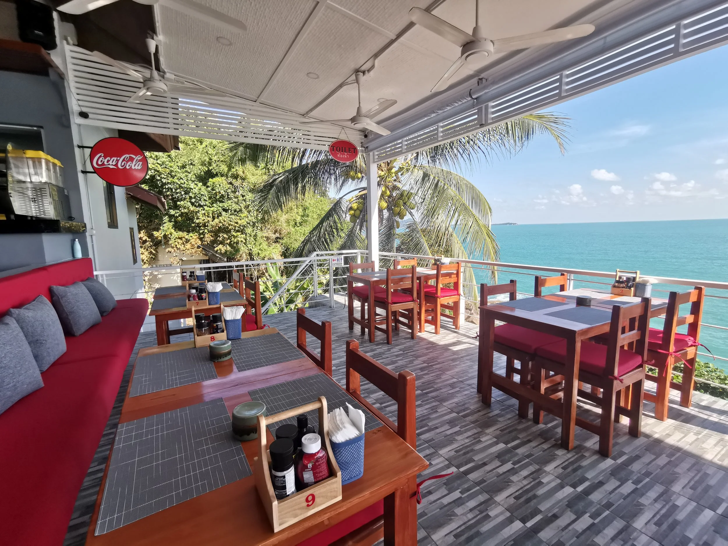 An outdoor restaurant with wooden tables and chairs facing the ocean. The ceiling has white fans and a sign indicating the toilet. There are lush green trees and palm trees in the background, and the sky is blue with some clouds.