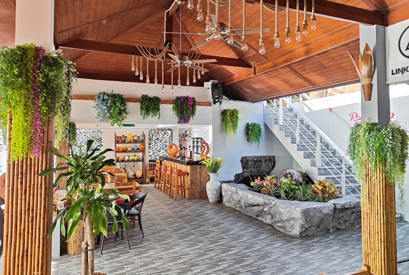 Indoor space with bamboo pillars, hanging plants, wooden ceiling with chandeliers, a bar area with stools, a seating area with a large plant, bookshelves, a water feature with plants, and a staircase.