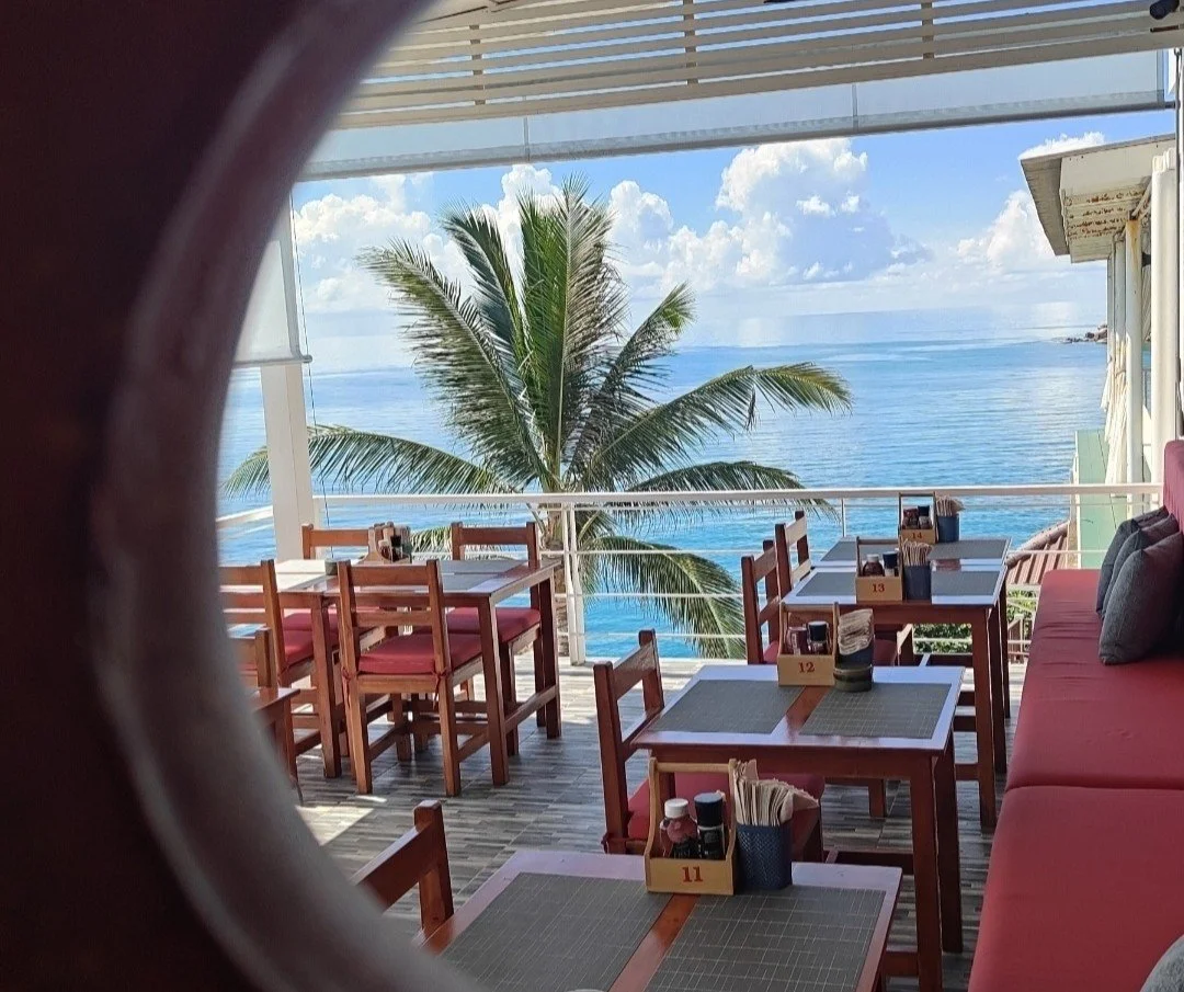 Empty outdoor restaurant with wooden tables and chairs, ocean view, palm tree, and partly cloudy skies.