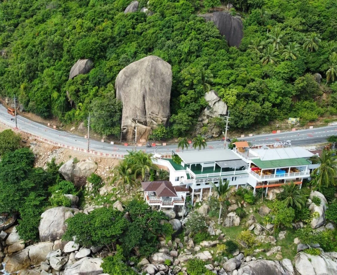A hillside with dense green trees and large rocks, a winding road passing through, and a white multi-level building with a patio and surrounding greenery at the base of the hill.
