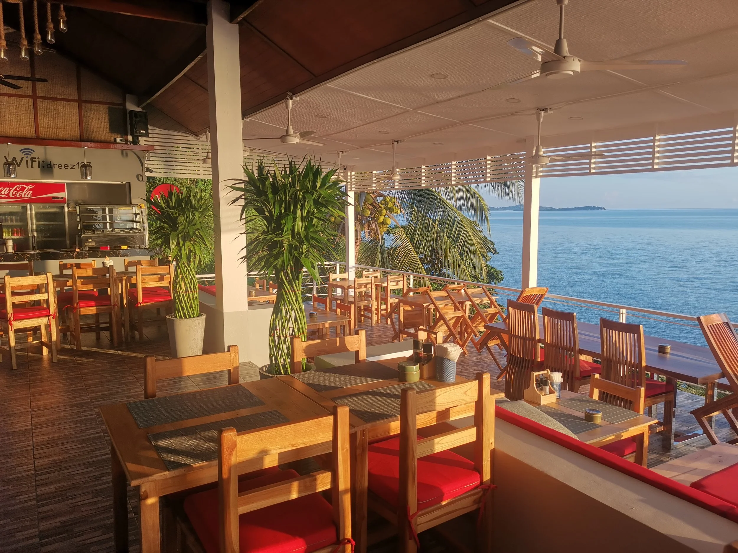 An indoor dining area with wooden tables and chairs with red cushions, overlooking an open balcony with a view of the ocean and palm trees, under a ceiling with ceiling fans.