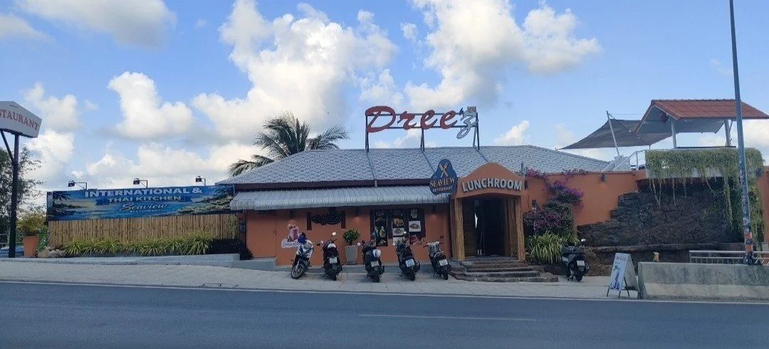 A restaurant on a street with five scooters parked in front, palm trees, and blue sky with clouds. The restaurant has a sign that reads "Druez" and another sign indicating a seafood and Thai cuisine.