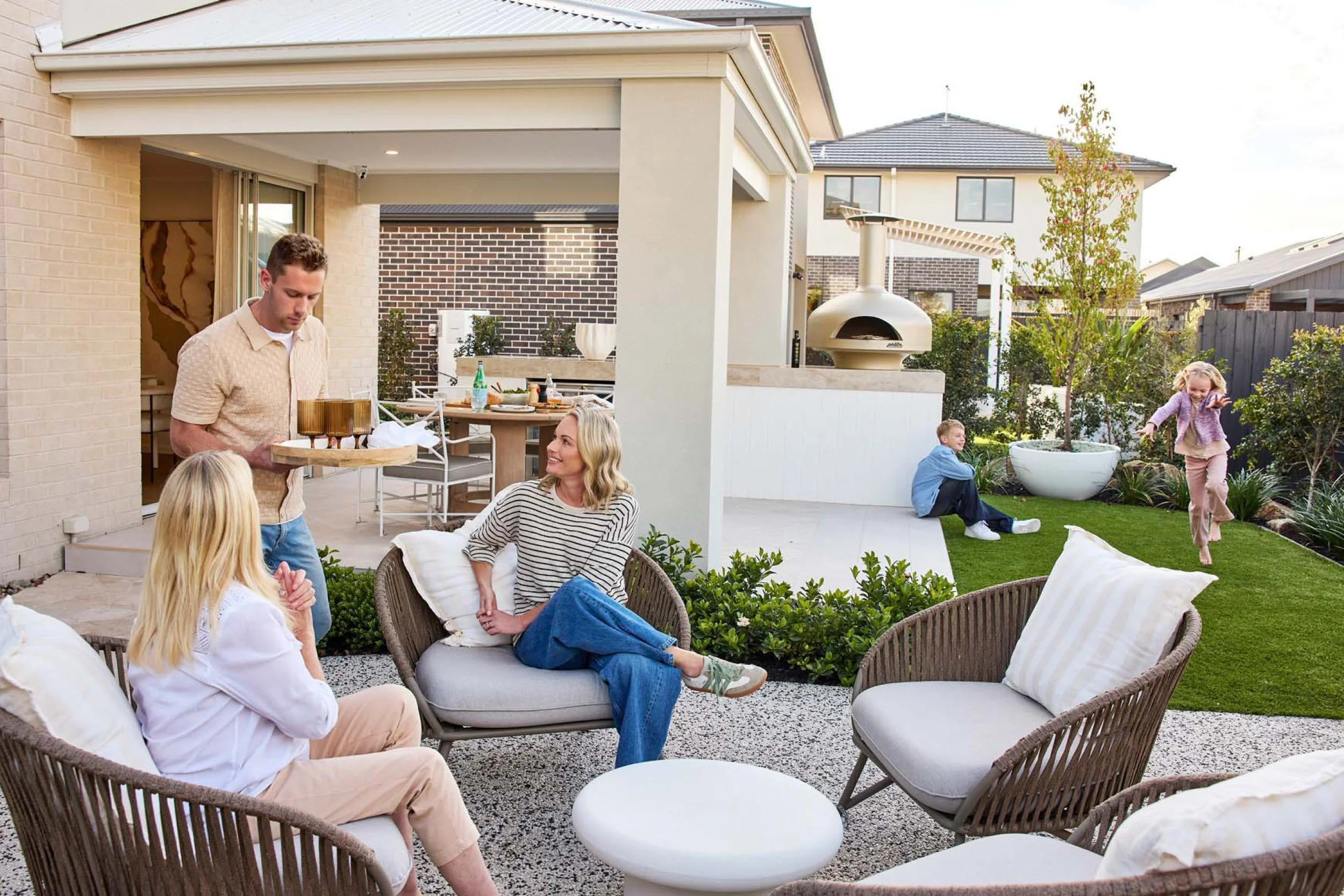 A family gathered outdoors in their backyard, enjoying a sunny day. A man is bringing food or drinks to a woman sitting on outdoor furniture, while a girl is playing and jumping on the grass, and a boy is sitting on the patio.