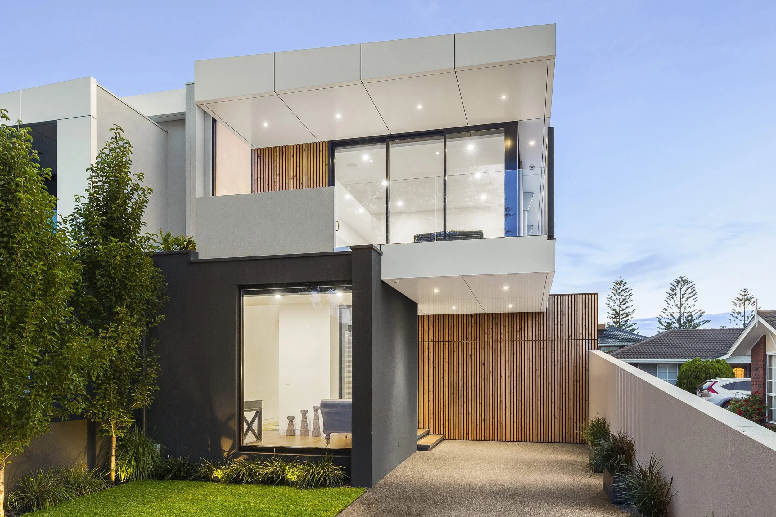 Modern two-story house with large glass windows, wooden paneling, and a well-maintained front yard with green grass and small plants, during dusk.