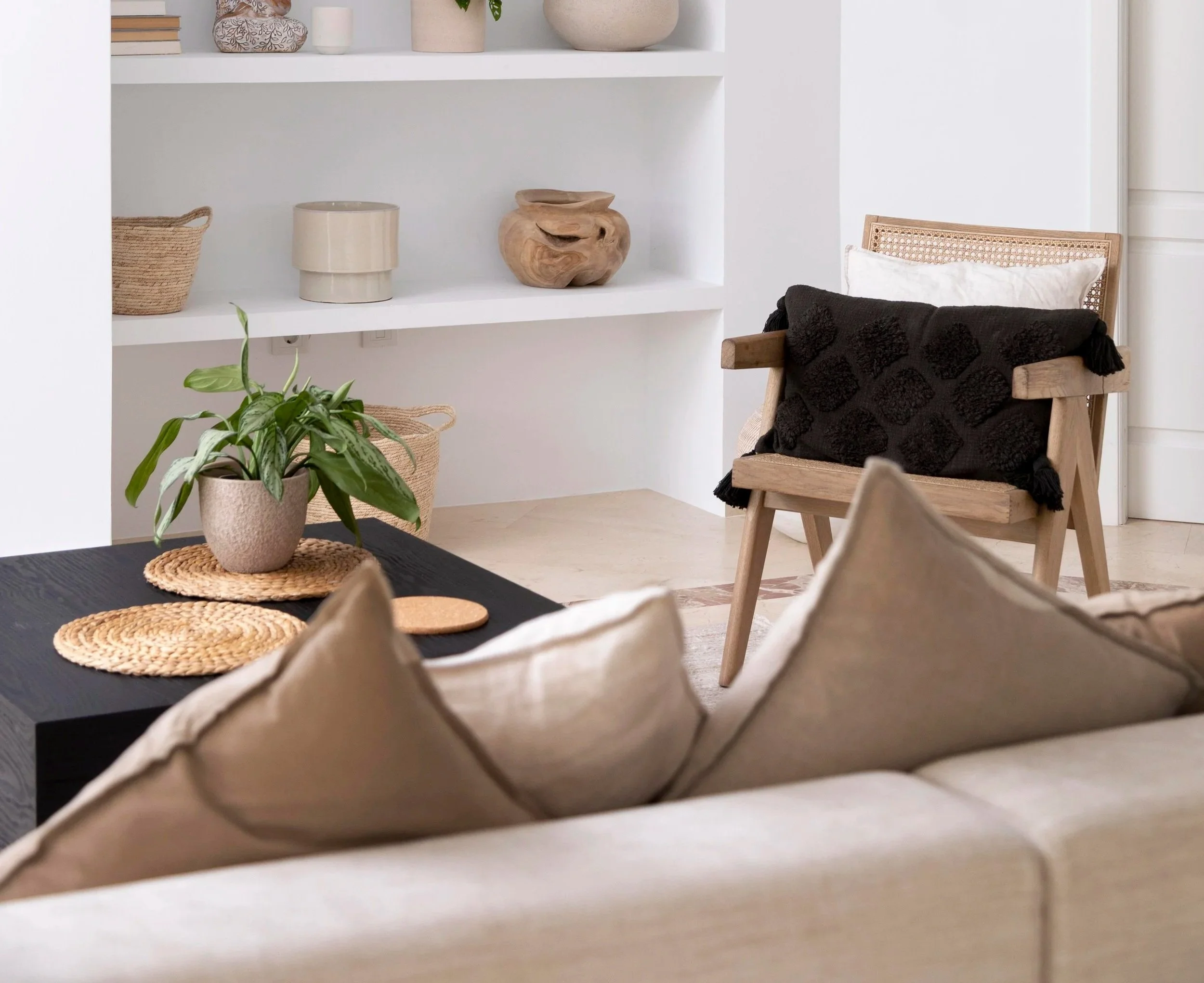 A cozy living room with beige sofa cushions, a black throw blanket on a wooden chair, a potted plant on a dark coffee table, and a white bookshelf with decorative items like vases and baskets.