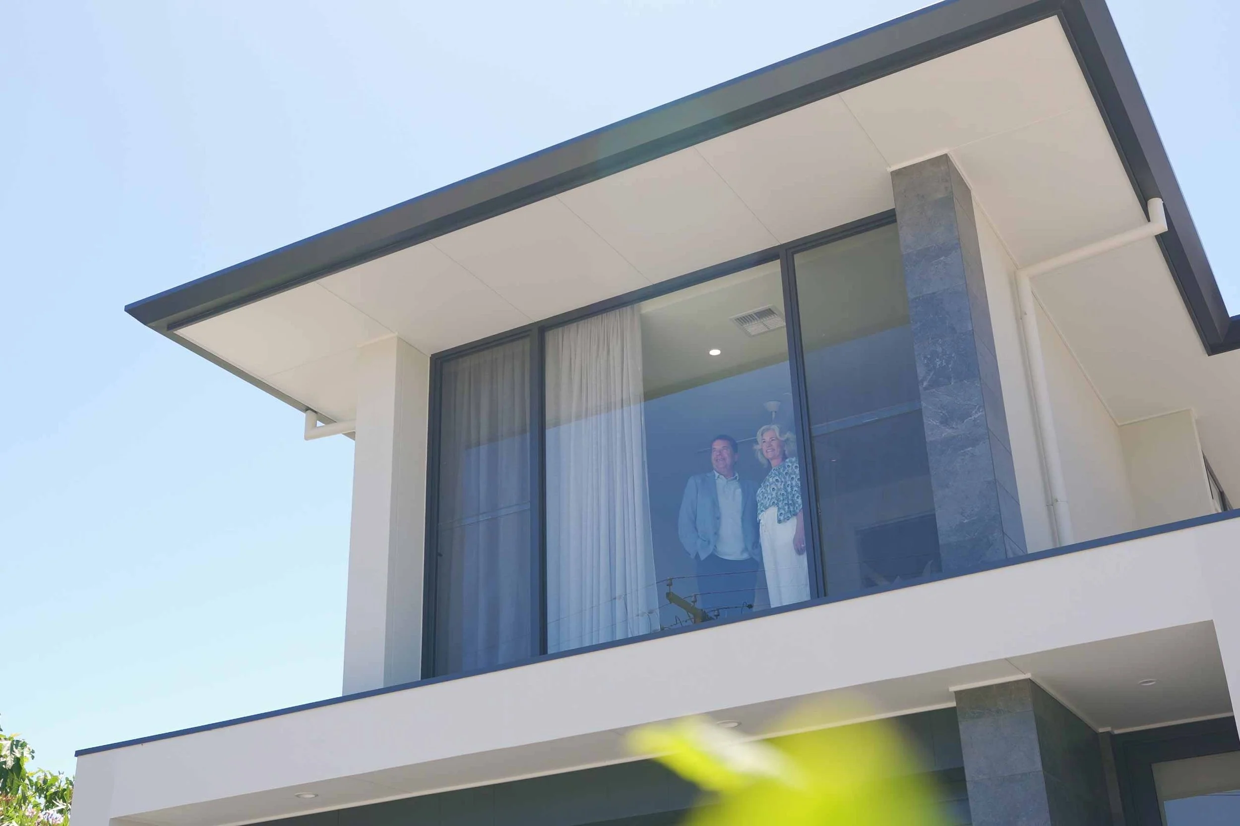 Two people standing inside a modern house looking out of a large glass window.