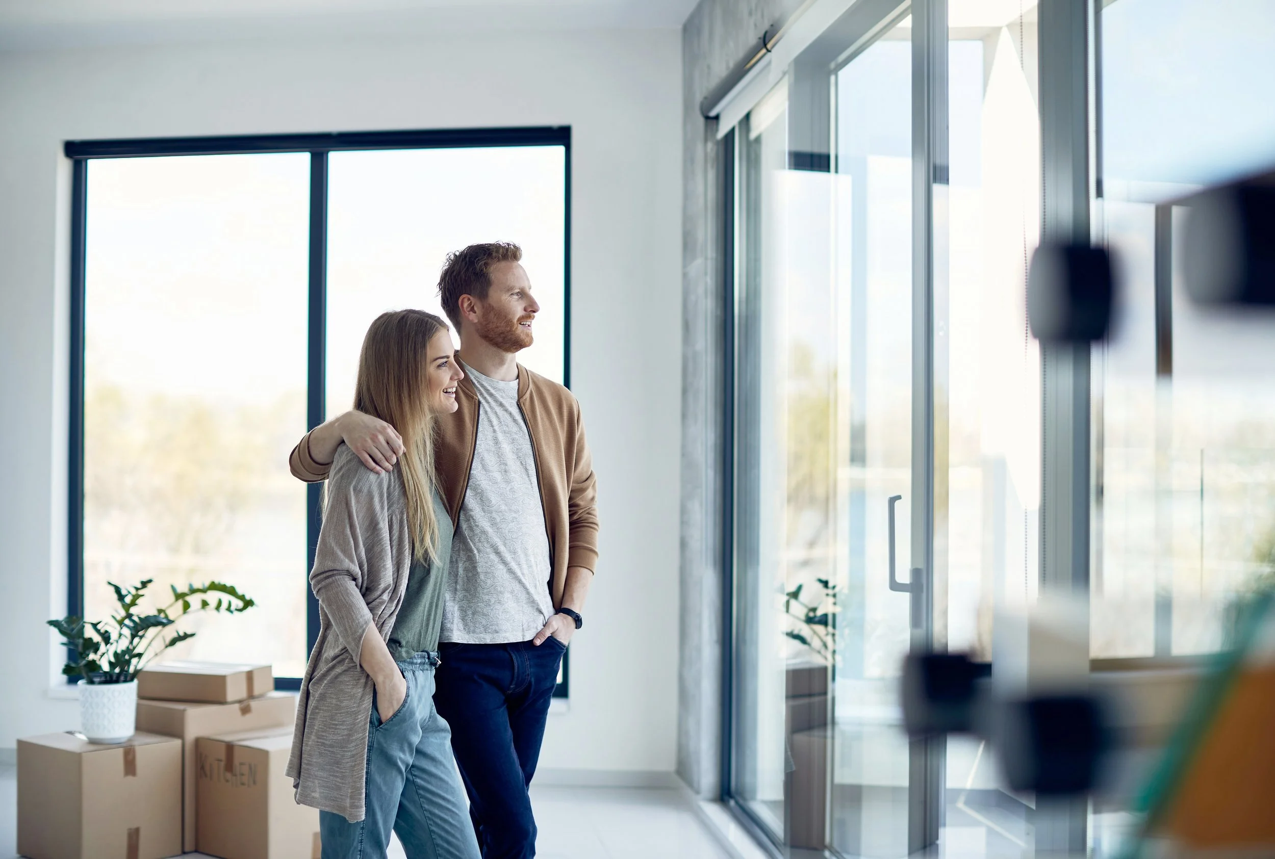 A young couple standing together in a bright, modern room, looking out the window with smiles.
