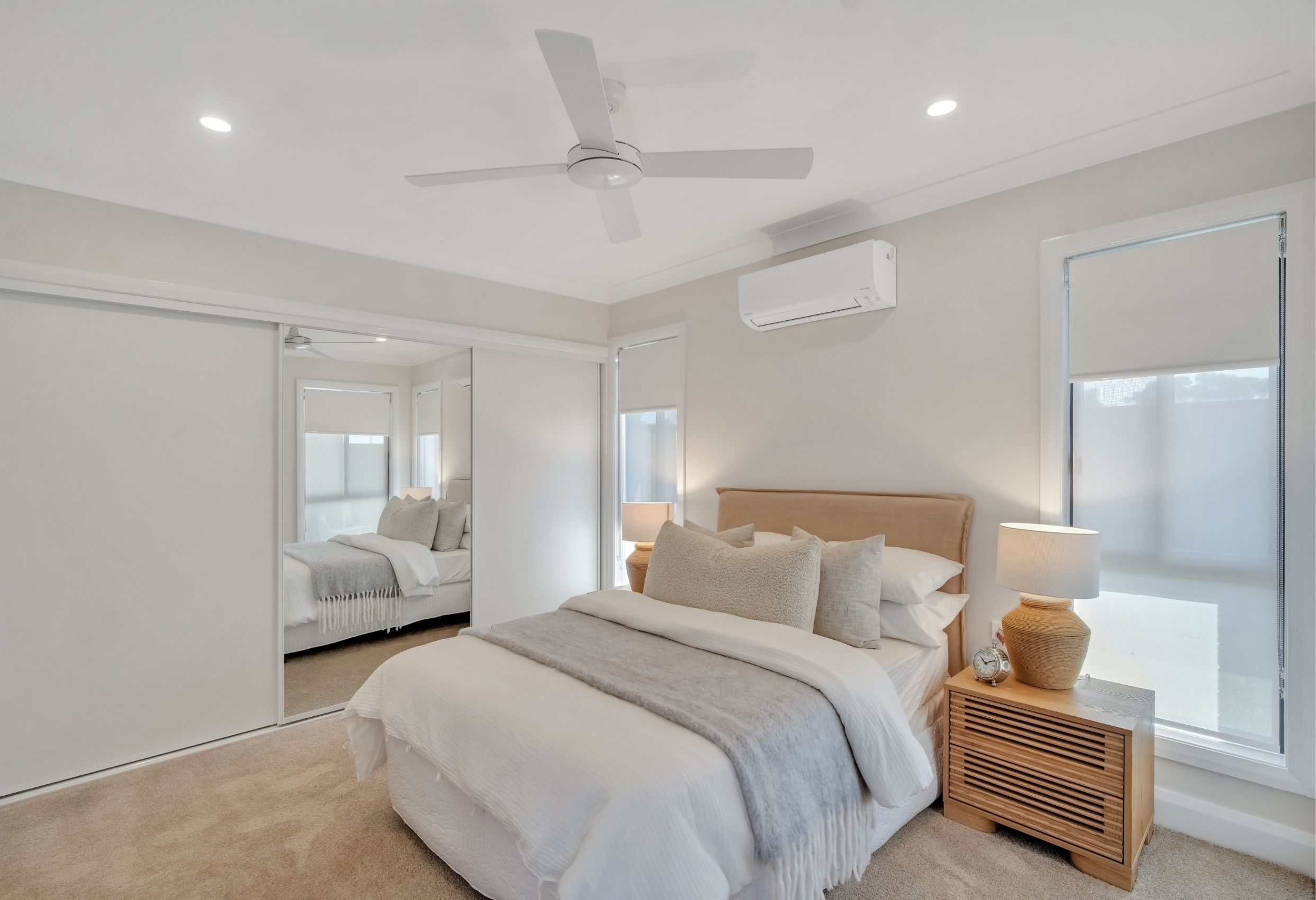A clean and bright bedroom with a beige bed frame, white bedding, and multiple pillows. There is a wooden nightstand with a lamp, clock, and some items. The room has two large windows with roller blinds, a ceiling fan, and an air conditioning unit. A mirrored sliding door closet reflects the bed.
