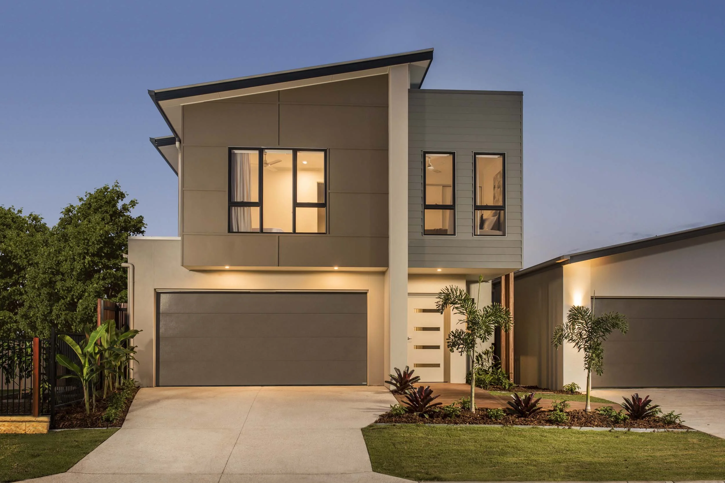 Modern two-story house with gray garage doors, a large front window with curtains on the upper floor, and a small landscaped front yard with tropical plants, during evening hours.