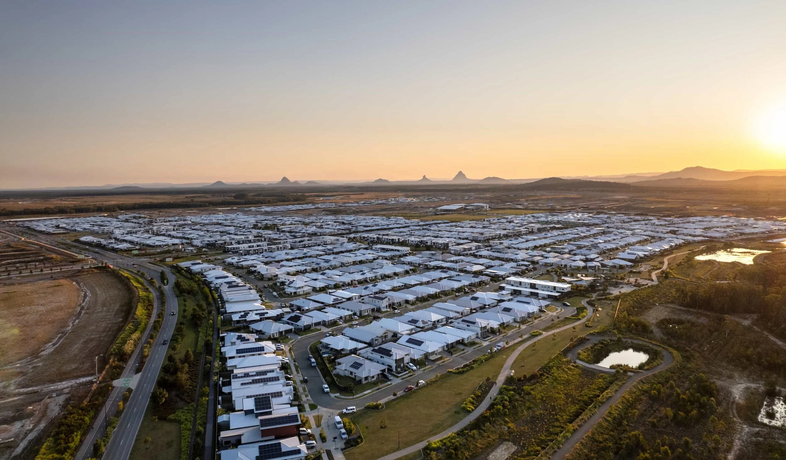 An aerial view of a suburban residential neighborhood at sunset, showing rows of houses with white roofs, roads, a park with a pond, and a vast open landscape in the distance.