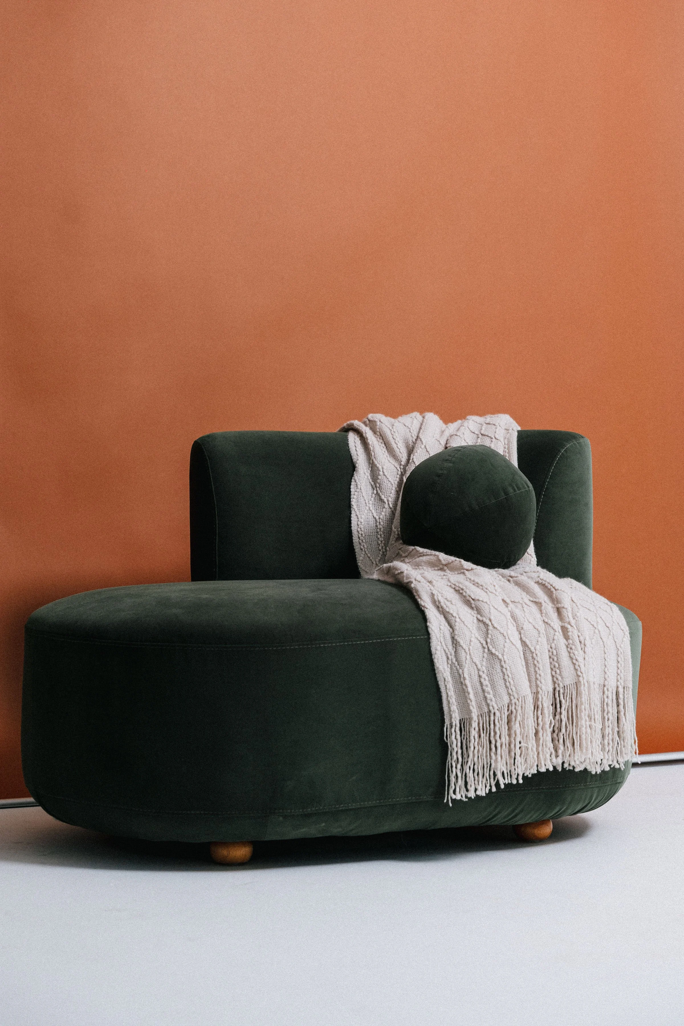 A dark green velvet armchair with a cream knitting throw draped over it and a round pillow resting on top, set against a warm terracotta wall.