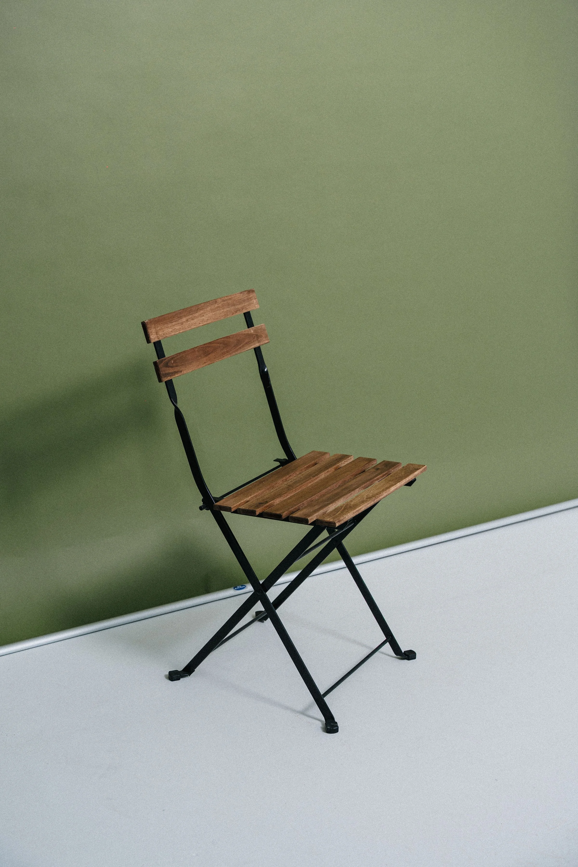 Wooden and metal folding chair with slatted seat and backrest, placed against a green wall and a light-colored floor.