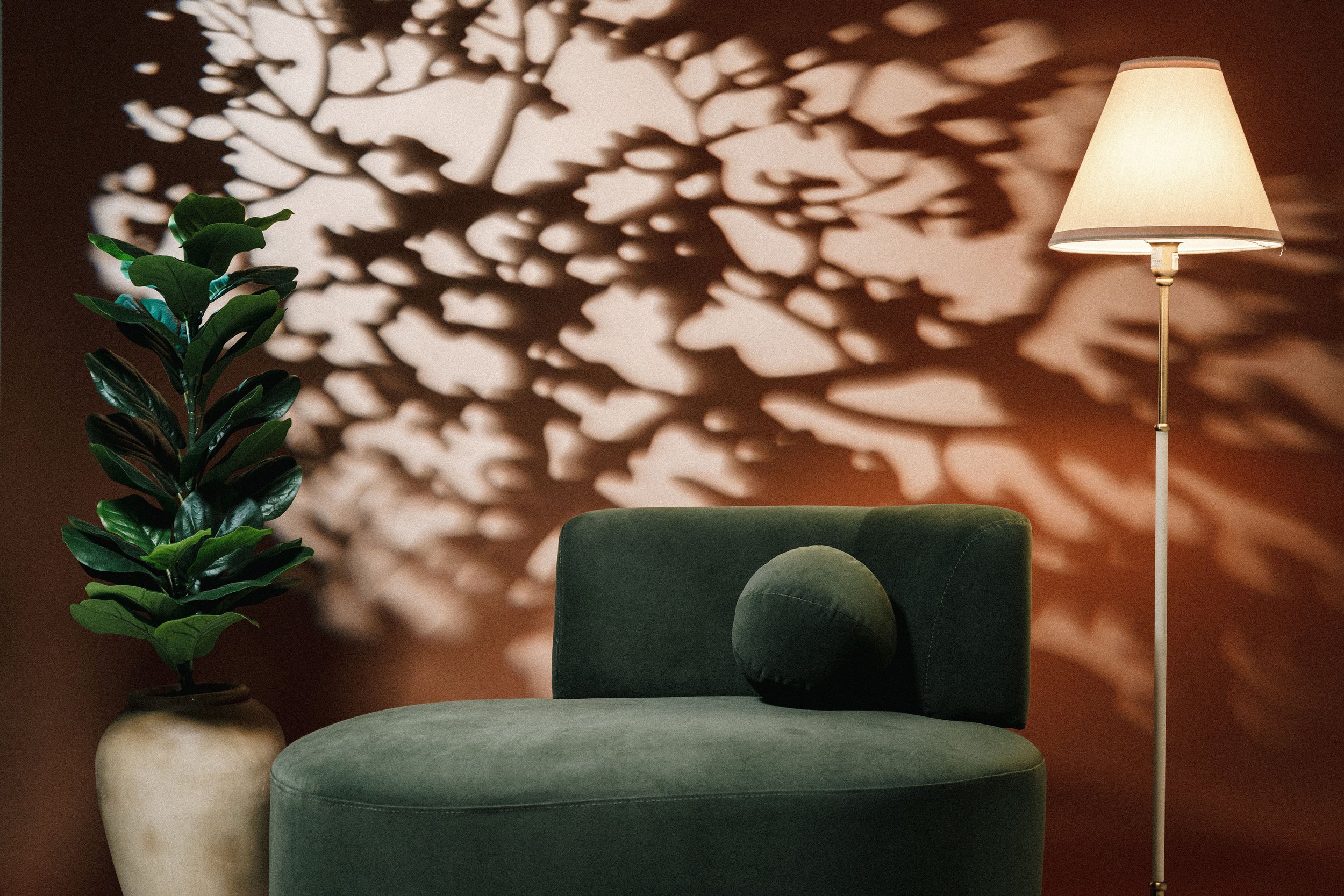 A green velvet chair with a round back pillow next to a tall lamp and a potted plant, with shadows of leaves cast on a brown wall in the background.
