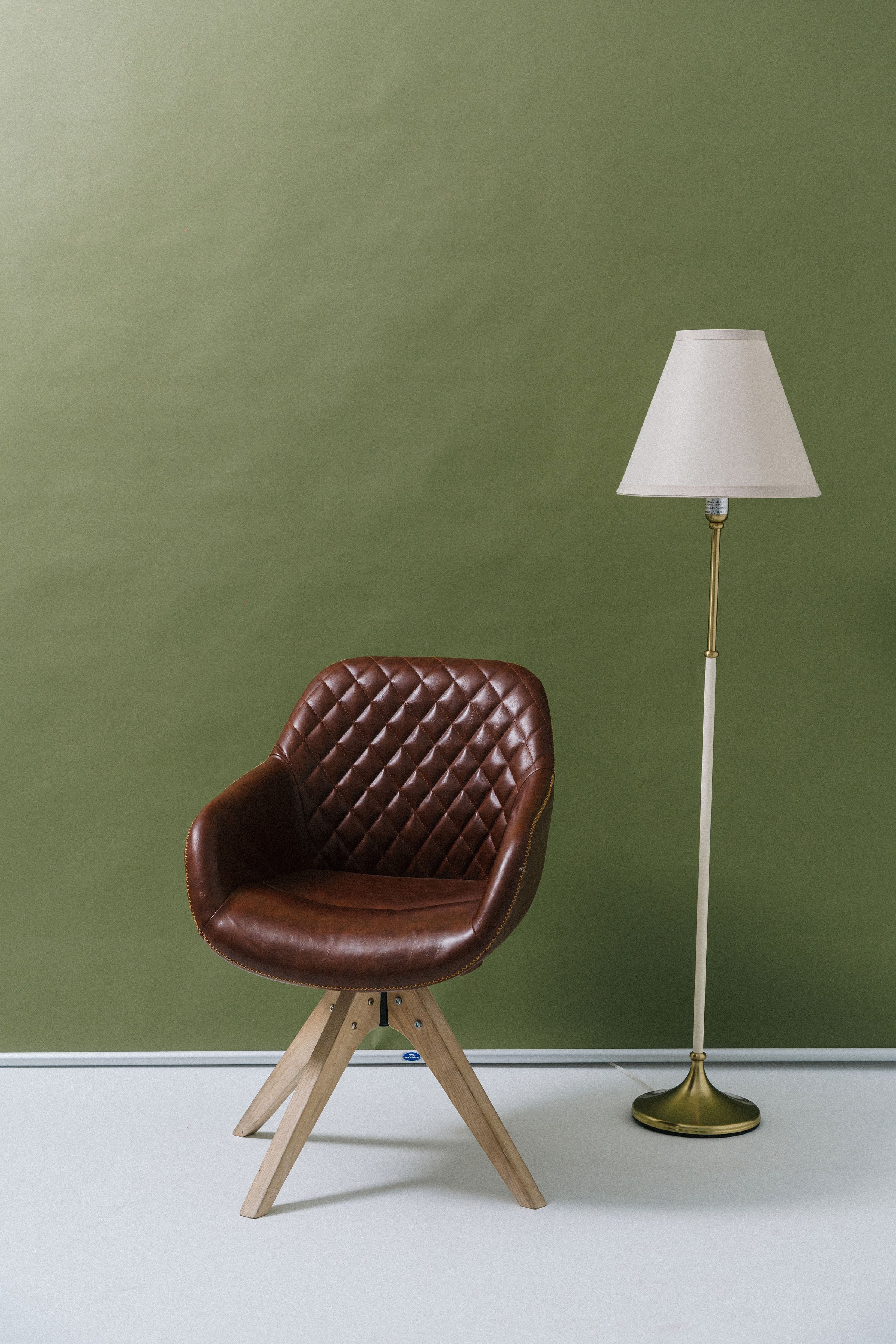 A brown leather chair with a quilted pattern on the backrest, placed next to a white and gold floor lamp against a green wall.