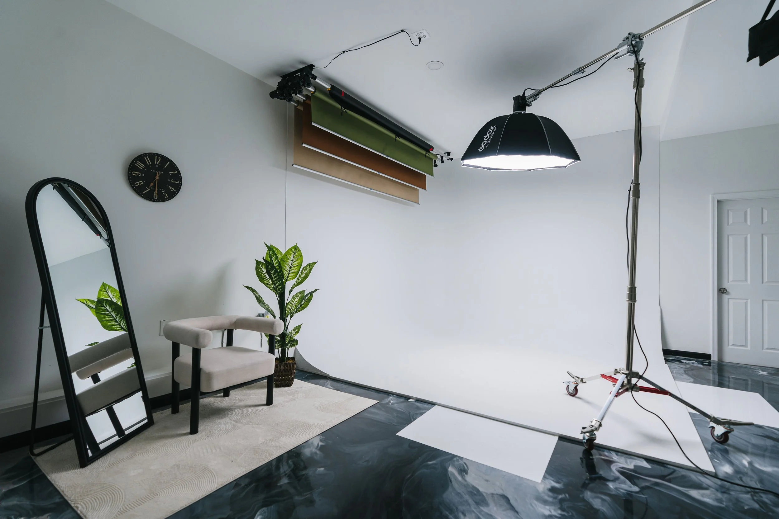 Photography studio with a white backdrop, professional lighting, a black-framed mirror leaning against the wall, a beige armchair, and a large indoor plant, with color swatches hanging from the ceiling.