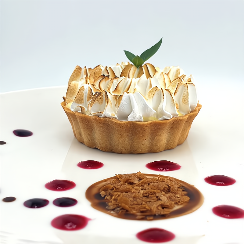 A lemon tart with toasted meringue topping and a sprig of basil, served on a white plate with red and purple sauce dots and a caramelized sugar disc.