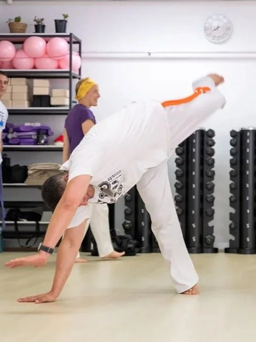 Person practicing capoeira in a fitness studio with two women in the background.
