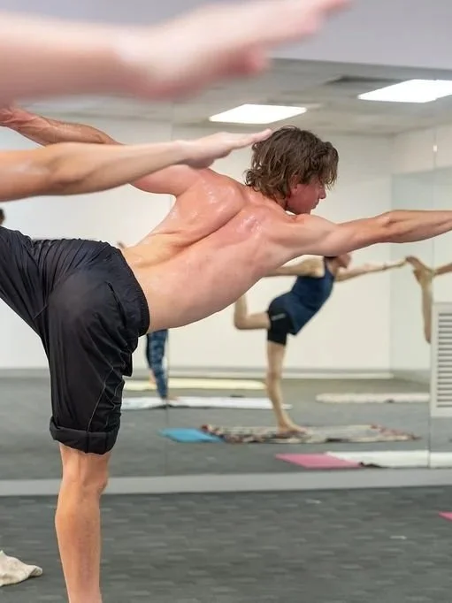 A man participating in a yoga class, balancing on one leg with arms extended, in a room with mirrors and other people practicing yoga in the background.
