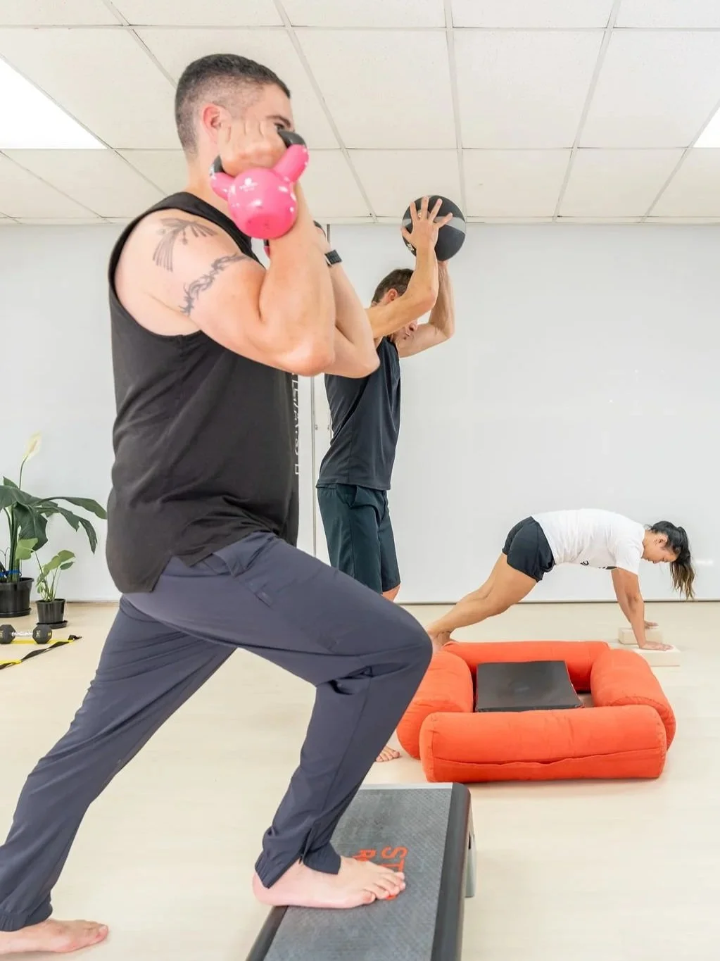 People exercising indoors, including a man lifting a kettlebell, a person holding a weighted ball and a woman in a plank position