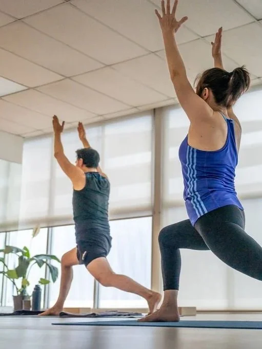 Two people practicing yoga in a bright studio. The woman in the foreground wears a blue tank top and black leggings, and the man in the background wears a dark tank top and shorts. Both are in a lunge pose with arms raised.