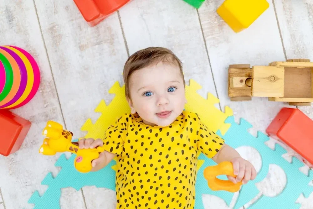 A baby in a yellow polka dot shirt lies on colorful foam mats, surrounded by vibrant toys, including blocks and a wooden truck, exuding a playful, joyful vibe.