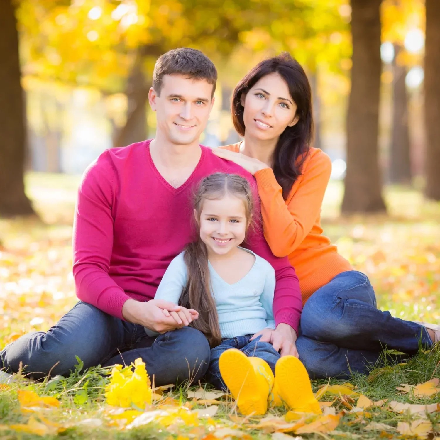 A smiling family sits on grass covered with autumn leaves. They are wearing casually colorful sweaters, with warm sunlight filtering through the trees.