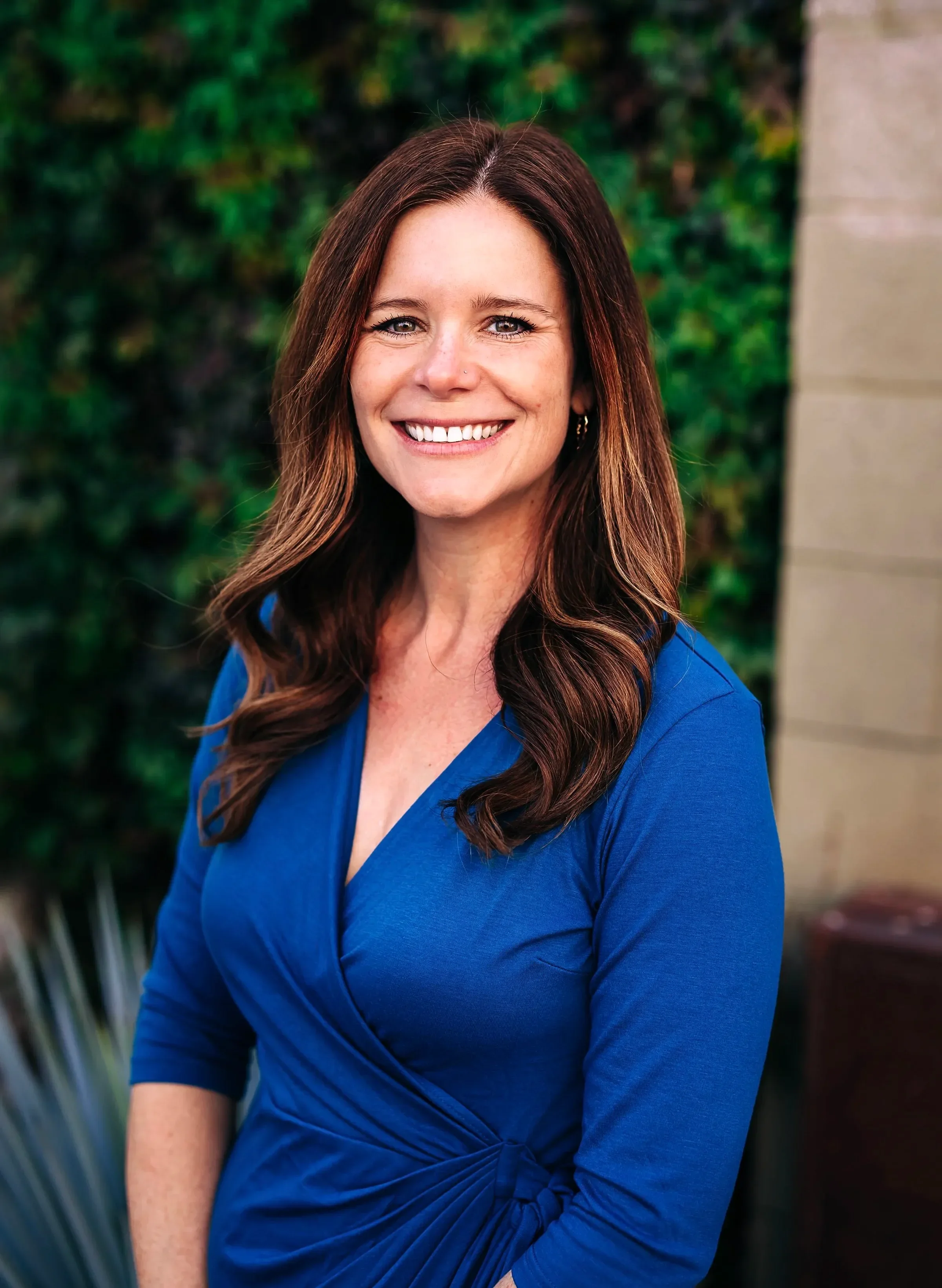 Dr. Wensman smiling, wearing a blue dress, stands in front of a leafy green wall. The image conveys warmth and friendliness.