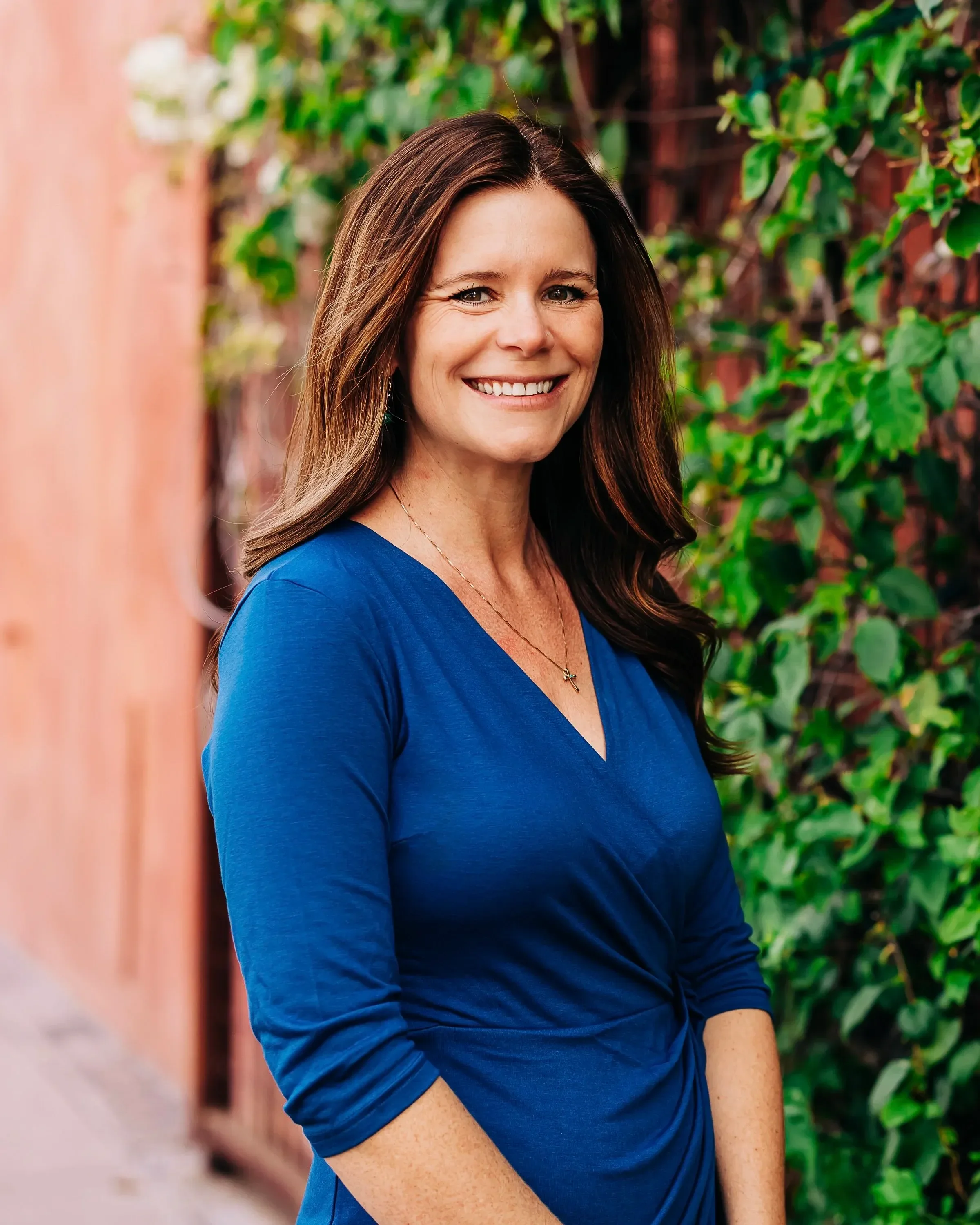 Dr. Wensman smiles warmly, standing against a backdrop of vibrant green ivy and a soft pink wall