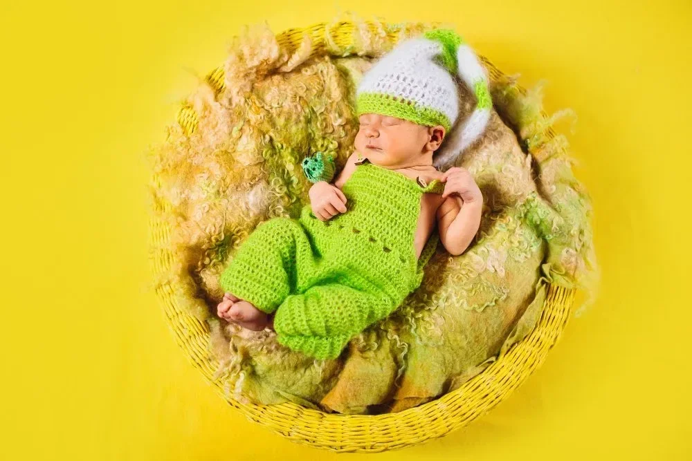 Sleeping baby dressed in a green crochet outfit and hat, lying in a yellow basket with soft textured blankets. Bright yellow background.