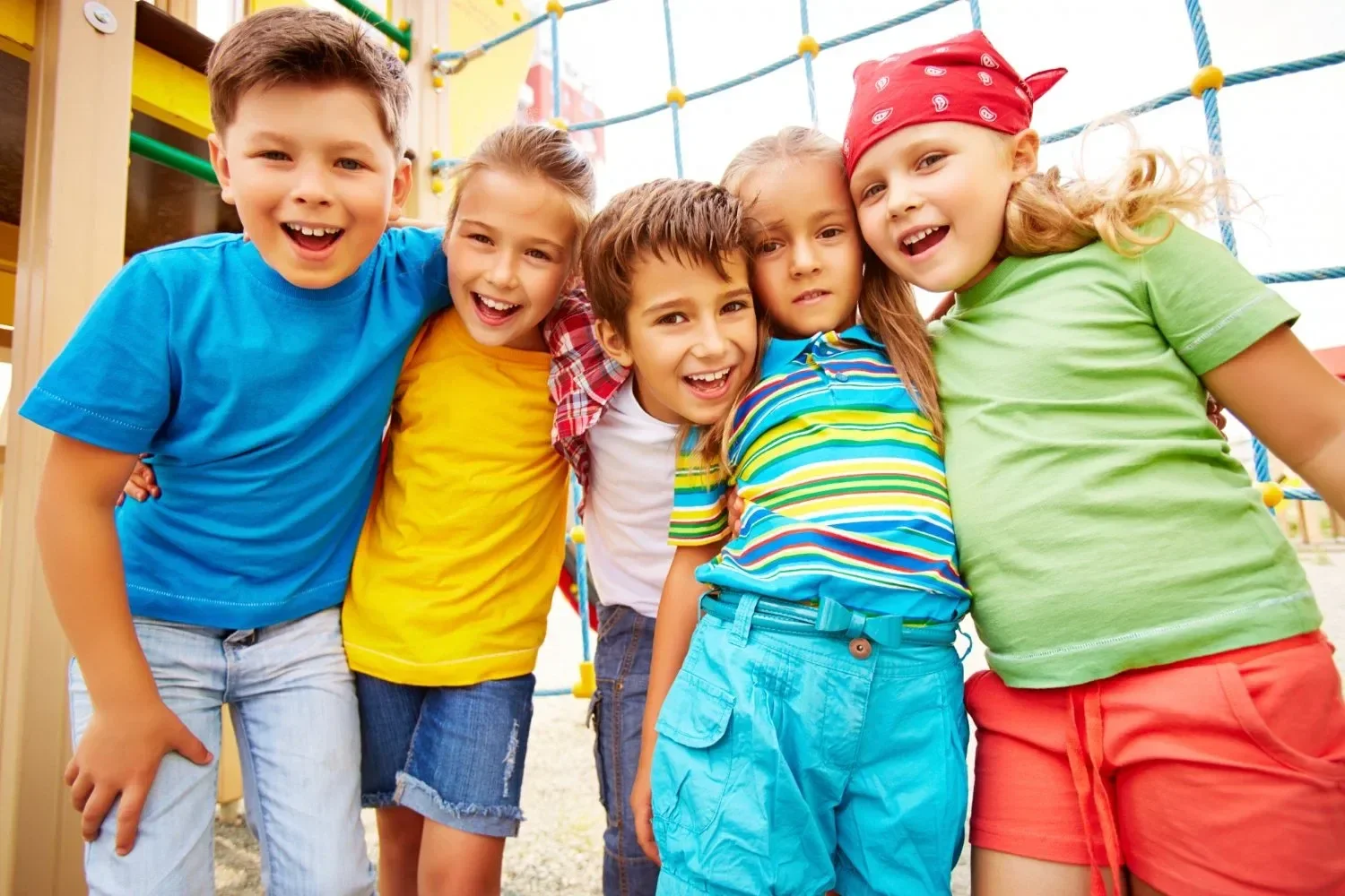 Five children in colorful clothing smile joyfully, hugging at a playground with climbing ropes. The scene conveys friendship and happiness.