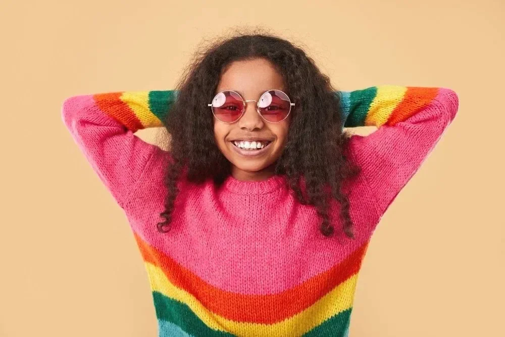 Smiling child in rainbow-striped sweater and pink sunglasses, arms behind head, against a beige background. Conveys a joyful, carefree vibe.