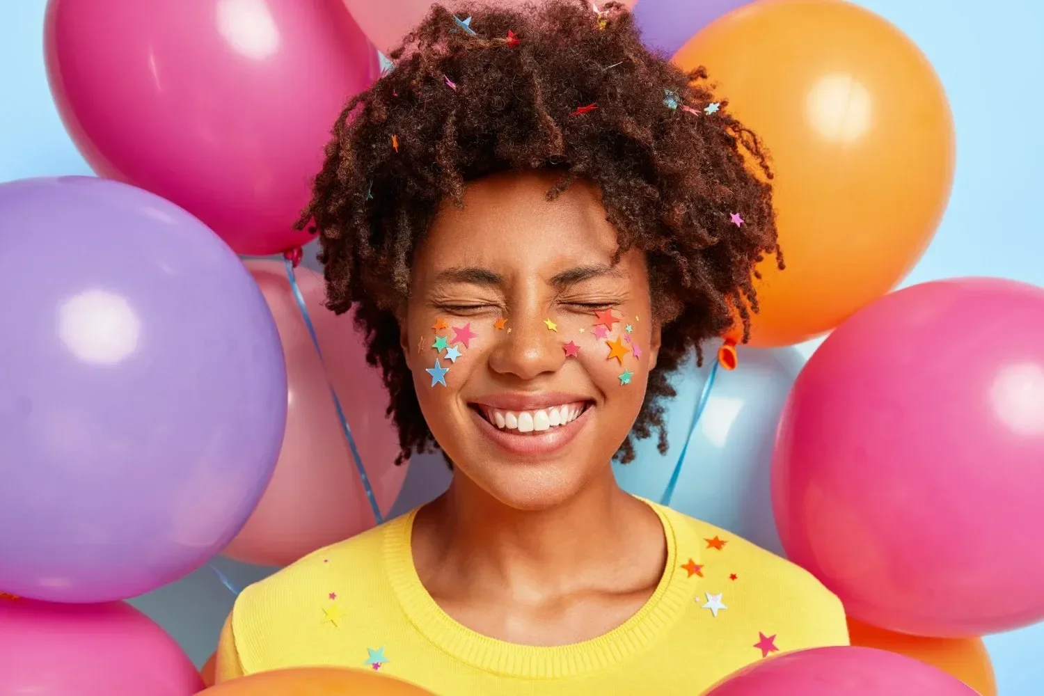 A joyful person with closed eyes and a big smile, surrounded by colorful balloons. Star confetti adorns their face, conveying a festive, cheerful vibe.