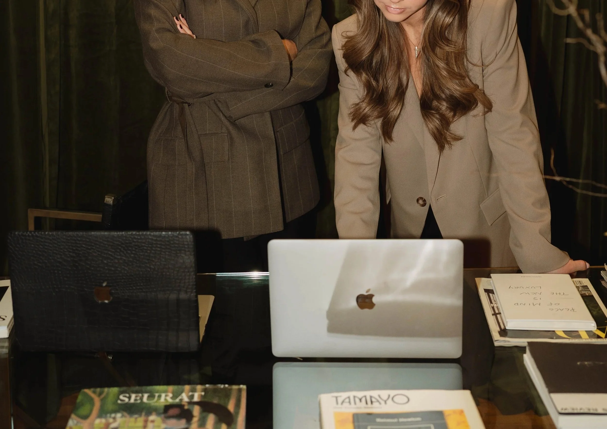 Two women in business attire standing behind a table with laptops, notebooks, and magazines, discussing or working together in an office setting.