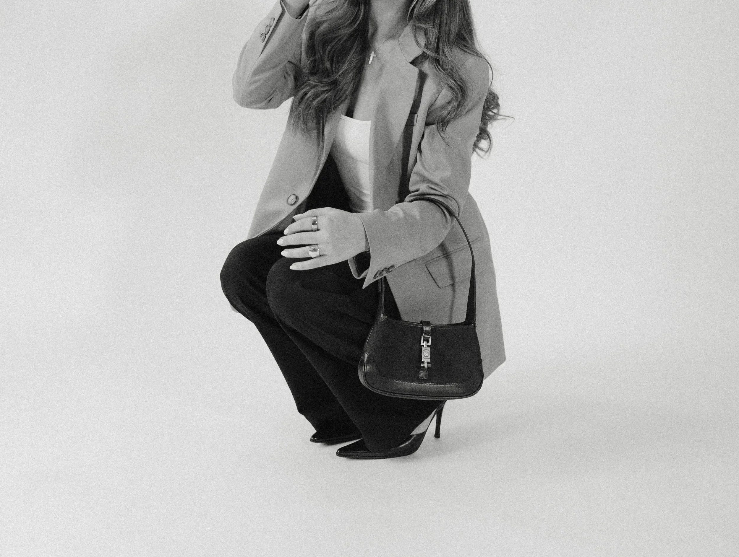 Black and white photo of a woman with long hair, wearing a blazer, cross necklace, rings, black high heels, and carrying a small black handbag, kneeling on one knee against a plain background.