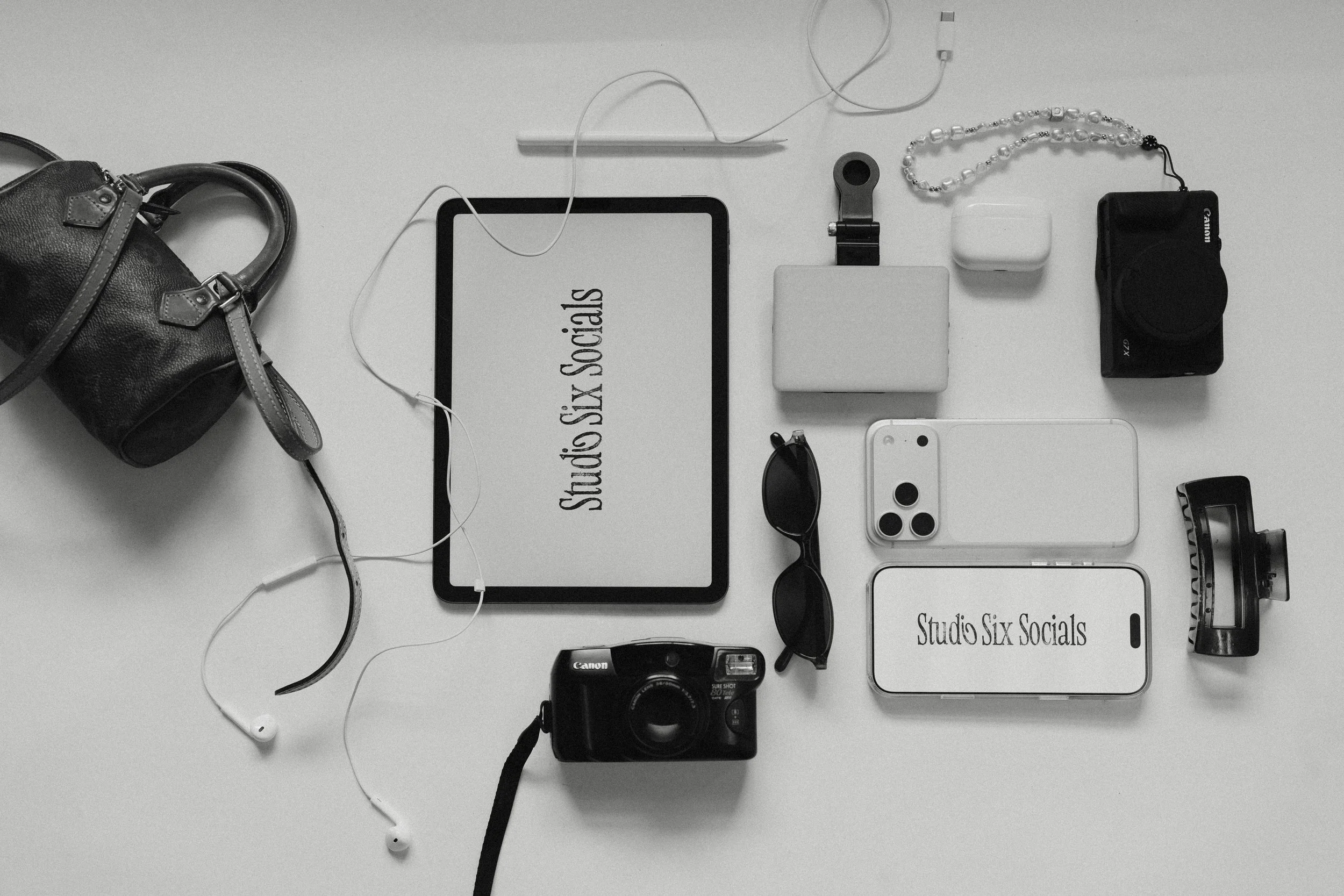 Flat lay of various electronic devices and accessories on a white surface, including a camera, smartphones, a tablet, wireless earbuds, sunglasses, a portable speaker, a power bank, and a small bag, with two screens displaying the text 'Studio Six Socials'.
