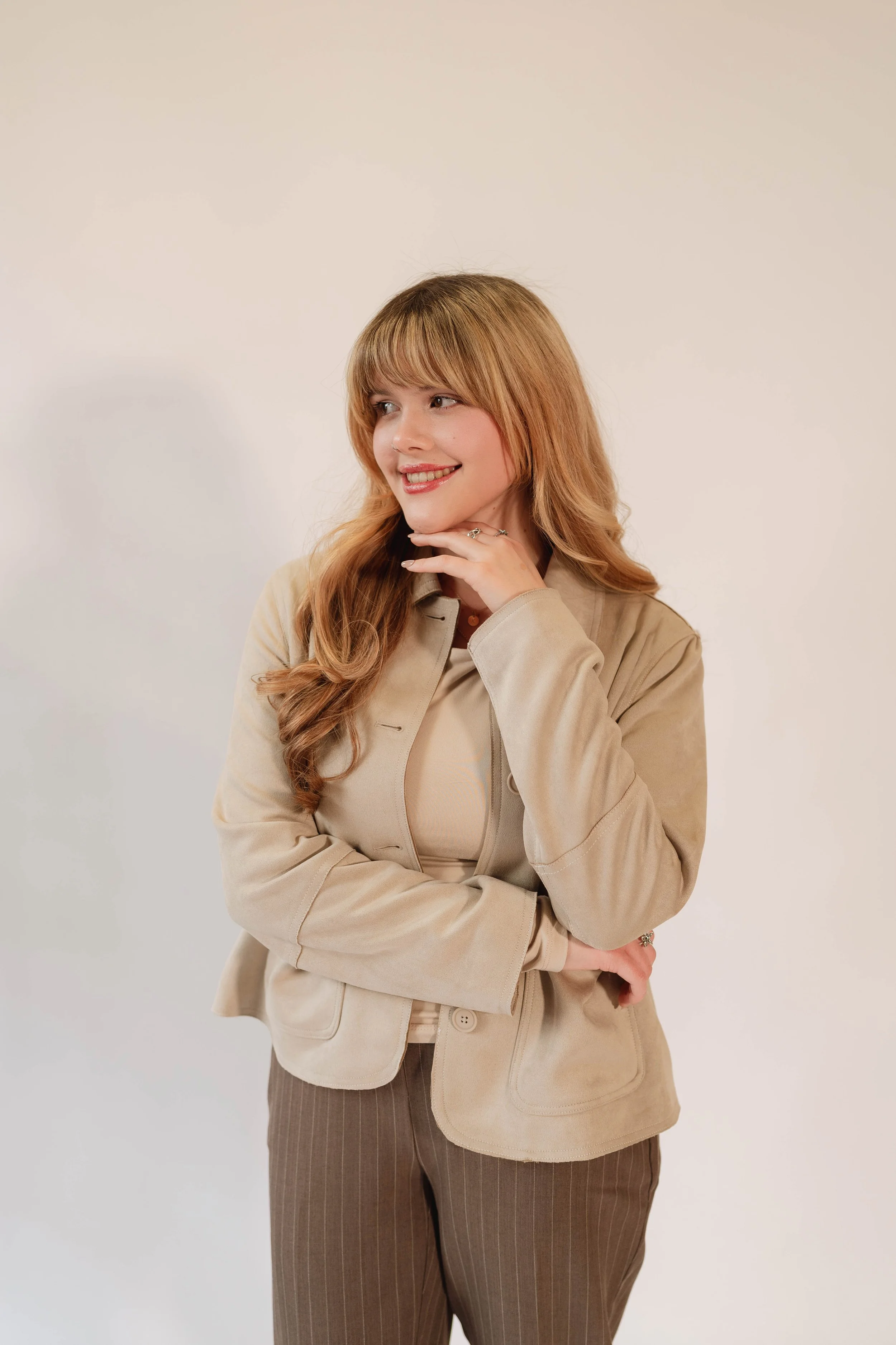 A woman with long, wavy blonde hair wearing a beige blazer and pinstripe trousers, smiling and posing with one hand touching her chin against a plain off-white background.