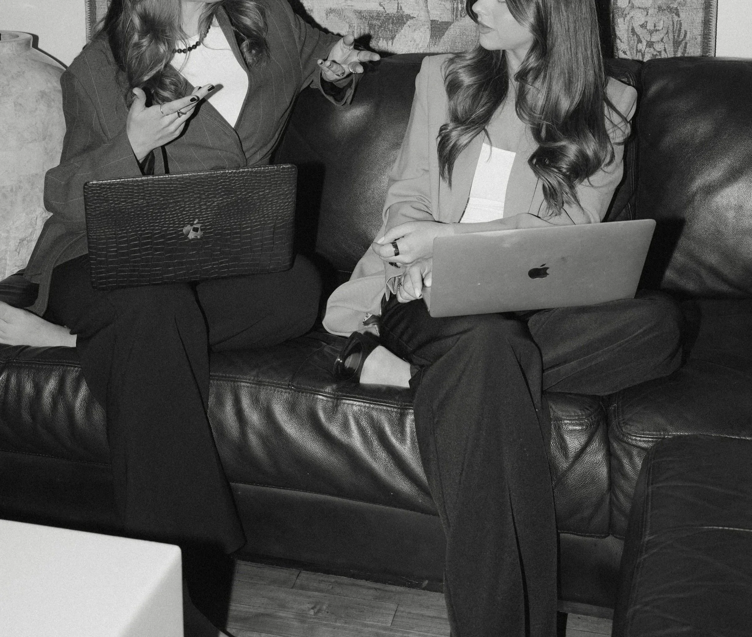 Two women sitting on a leather couch have laptops on their laps, engaging in conversation, with one woman gesturing.