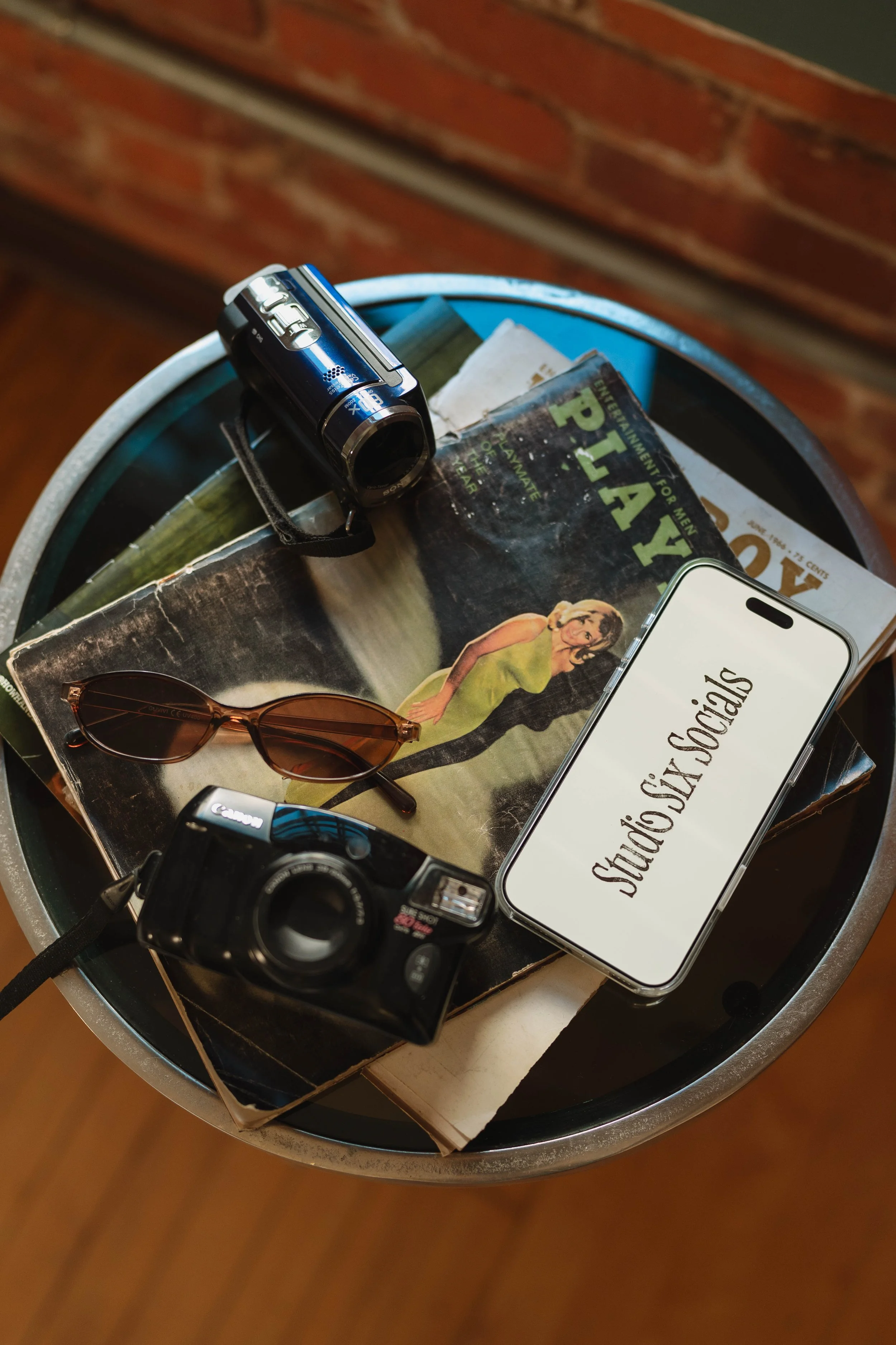 A round side table holding a vintage camera, sunglasses, a video camera, a phone with 'Studio Six Socials' displayed, and magazines, with a brick wall in the background.