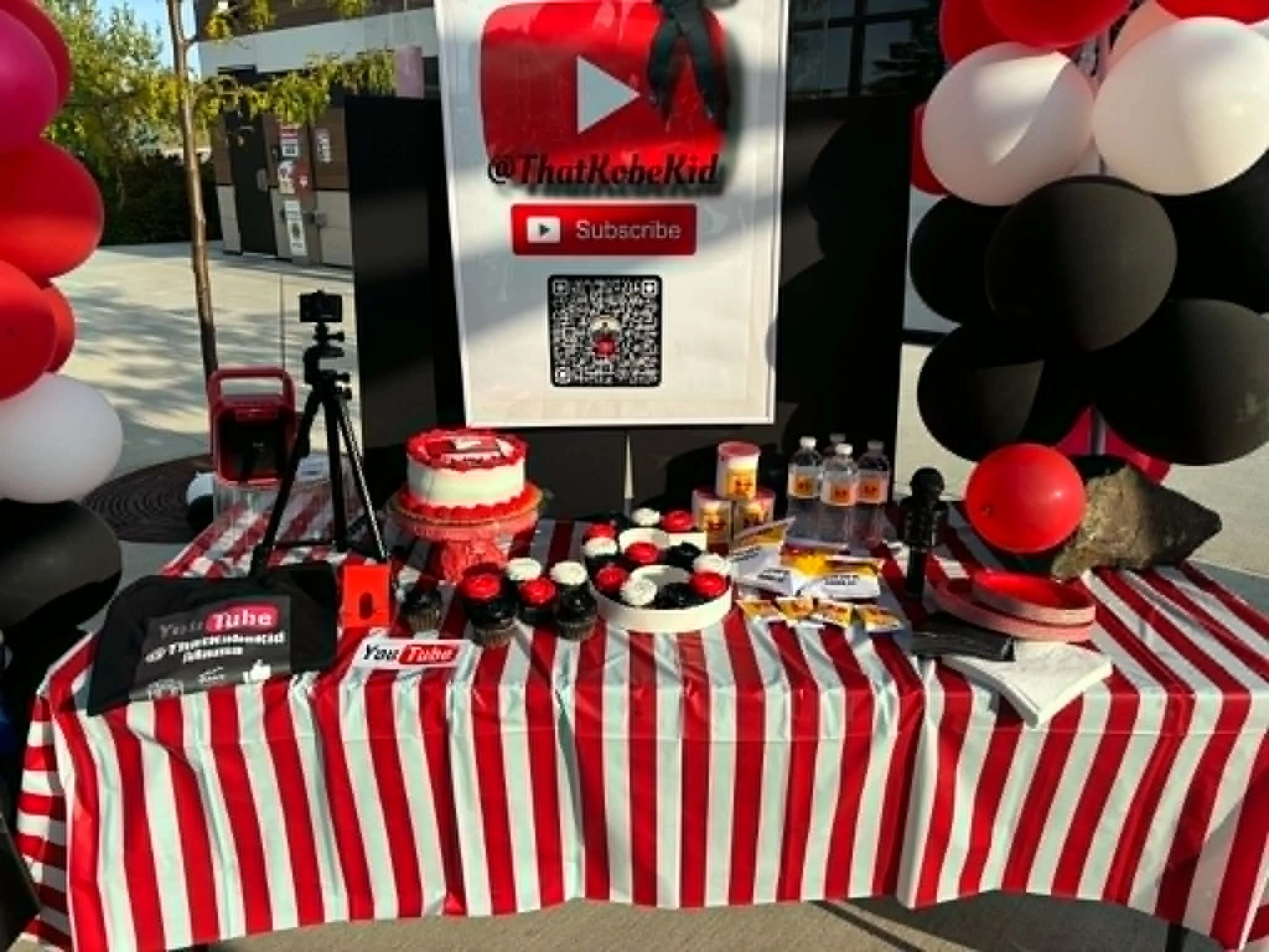 A table with a red, white, and black striped tablecloth featuring various items and decorations, including a cake with a red and white design, drinks, and numerous small containers, all arranged for a celebration or event. There are balloons surrounding the table, and a sign with a YouTube logo, a QR code, and social media information.