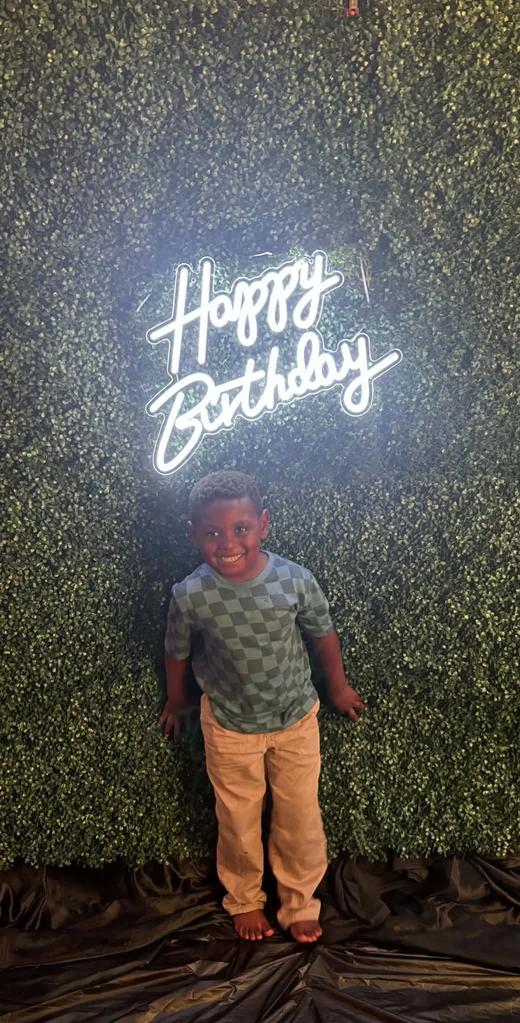 A young boy standing on black fabric in front of a green hedge wall with a neon sign that says 'Happy Birthday'. The boy is smiling, wearing a checkered green and gray T-shirt and khaki pants.