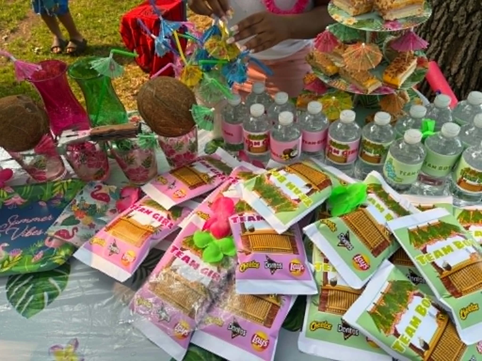 Table displaying various packs of party supplies including colorful paper umbrellas, bottled water, and plastic gift bags, outdoors on grass.