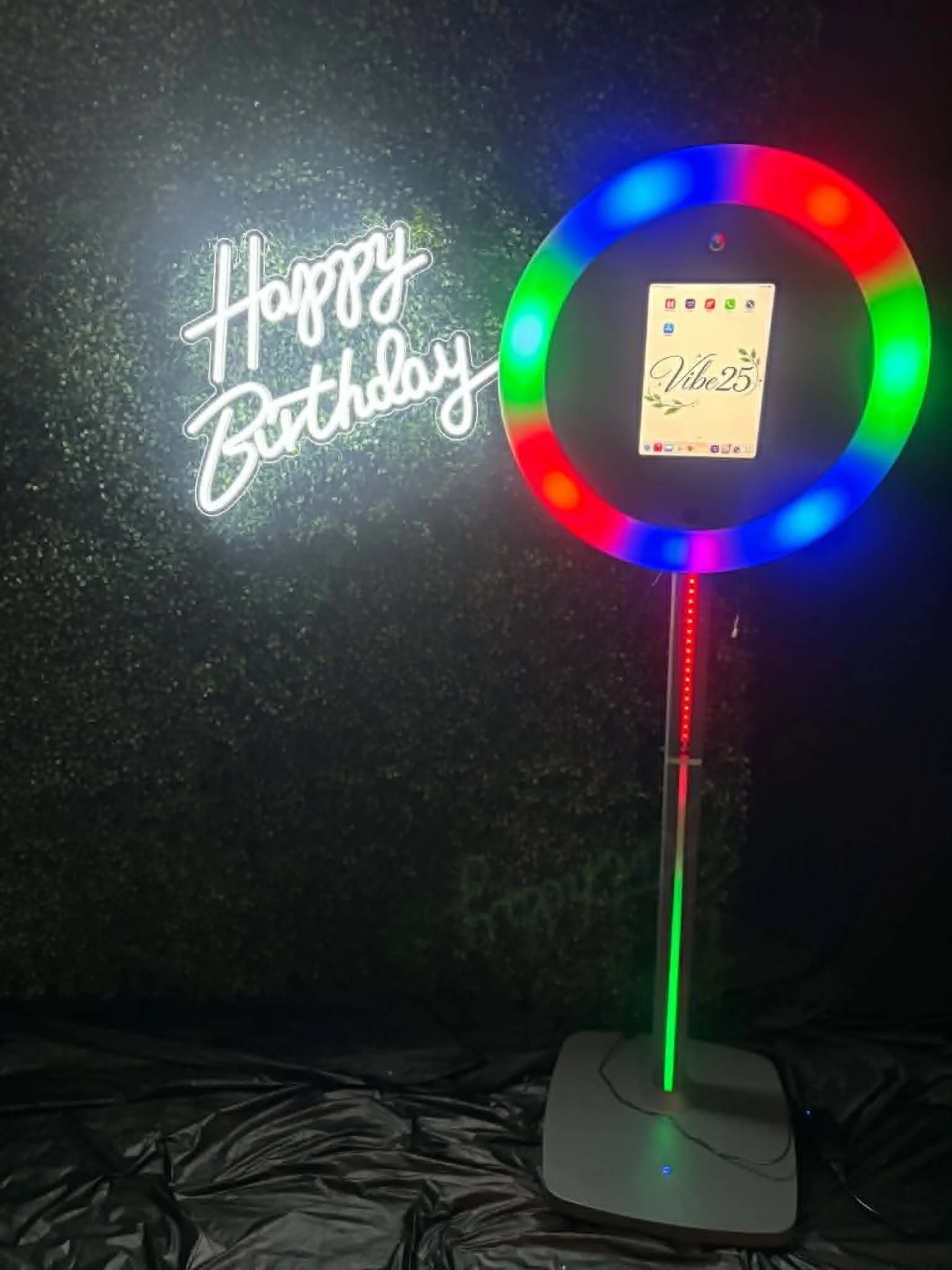A colorful LED rainbow light on a stand, placed on a surface protected with reflective foil, with a neon sign reading ‘Happy Birthday’ in the background.