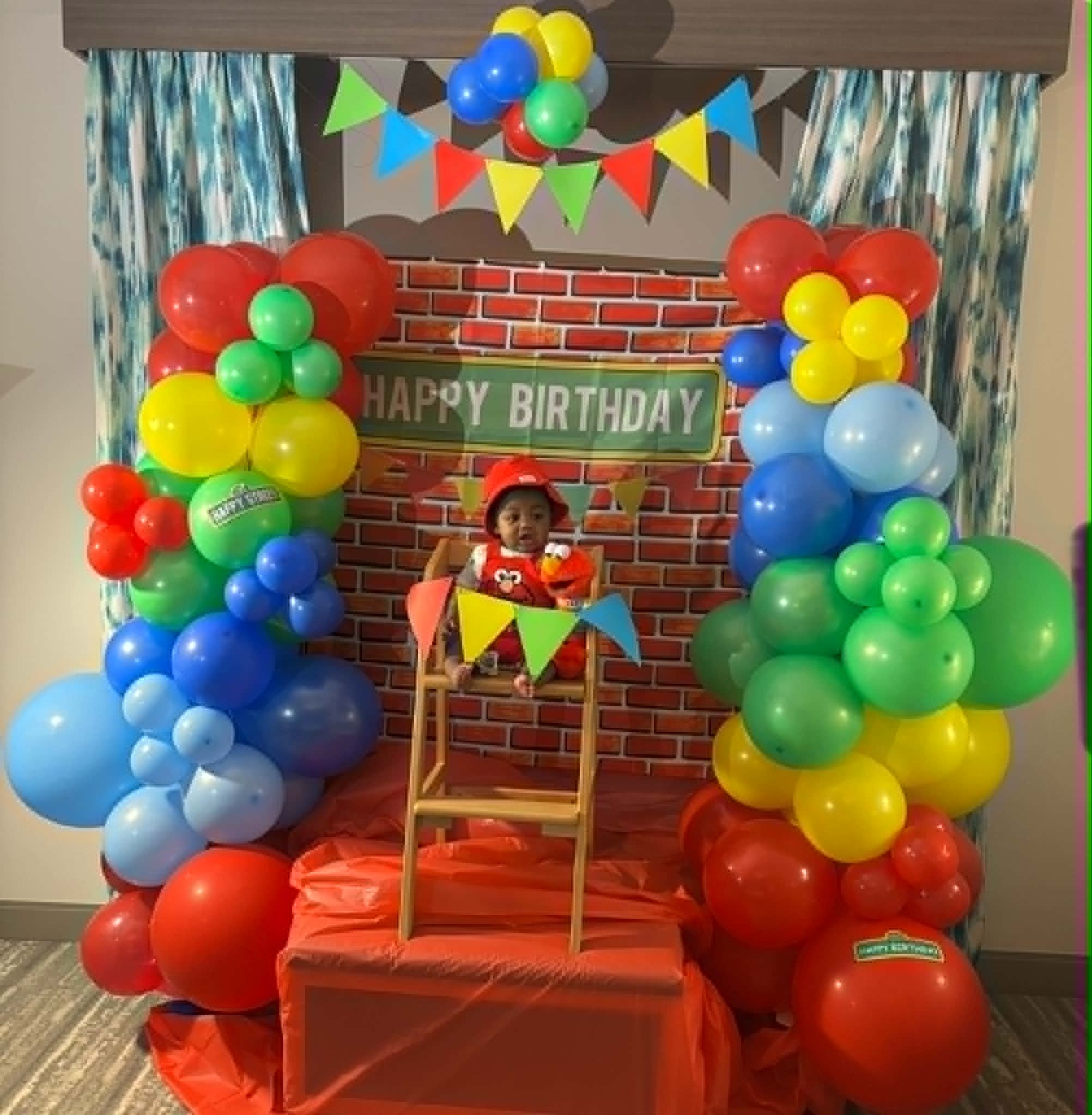 A colorful birthday party decoration with red, blue, yellow, green, and light blue balloons that form a border around a brick-patterned backdrop. The backdrop has a green banner that says 'HAPPY BIRTHDAY' and some bunting flags hanging across the top. There is a child wearing a red birthday crown, sitting on an orange chair, and some patterned curtains are visible on the sides.
