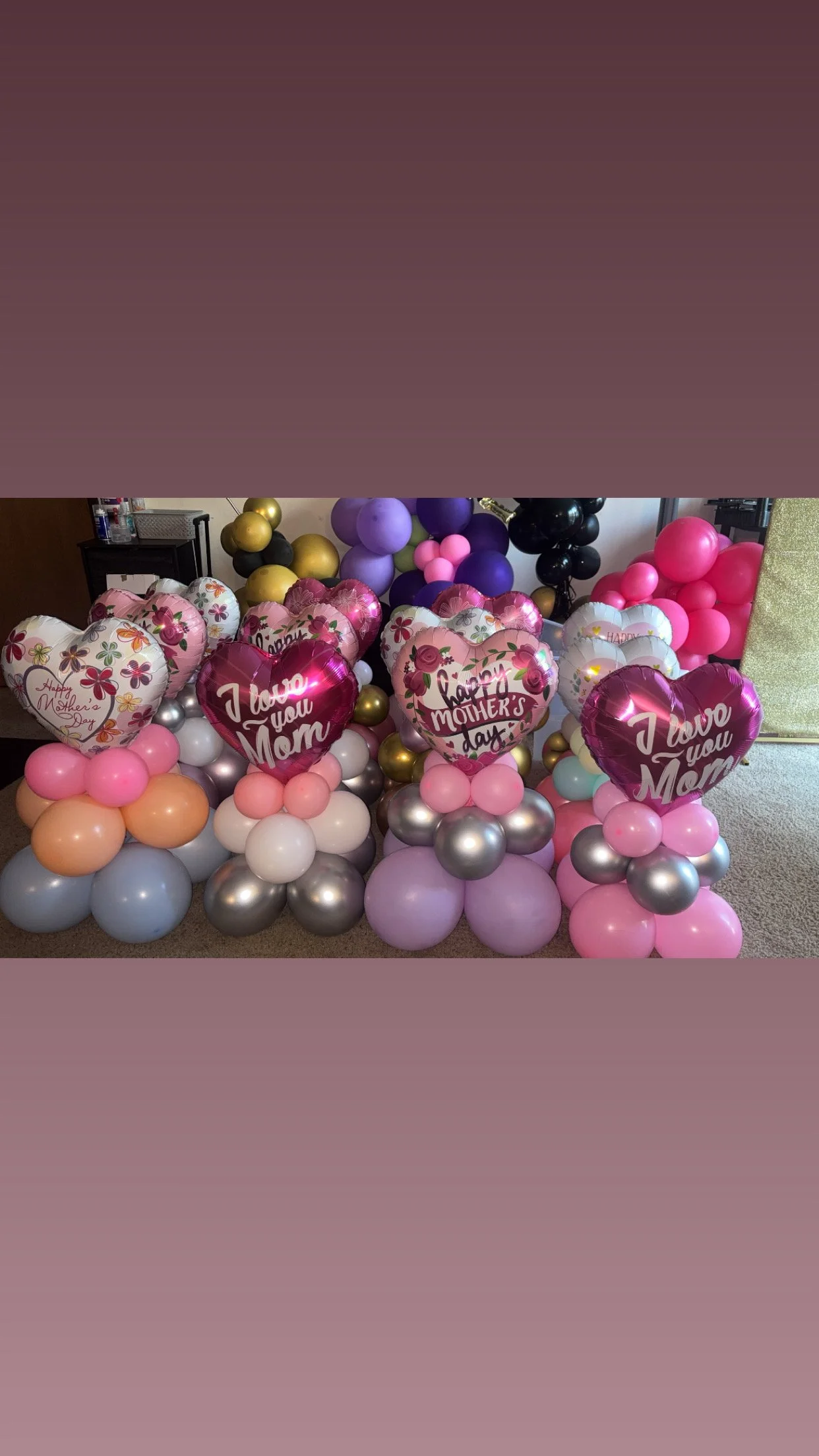 A collection of balloons arranged in clusters, with some shaped like hearts and others round, featuring messages such as 'I love you Mom' and 'Happy Mother's Day', in pink, purple, black, gold, silver, peach, and white colors.